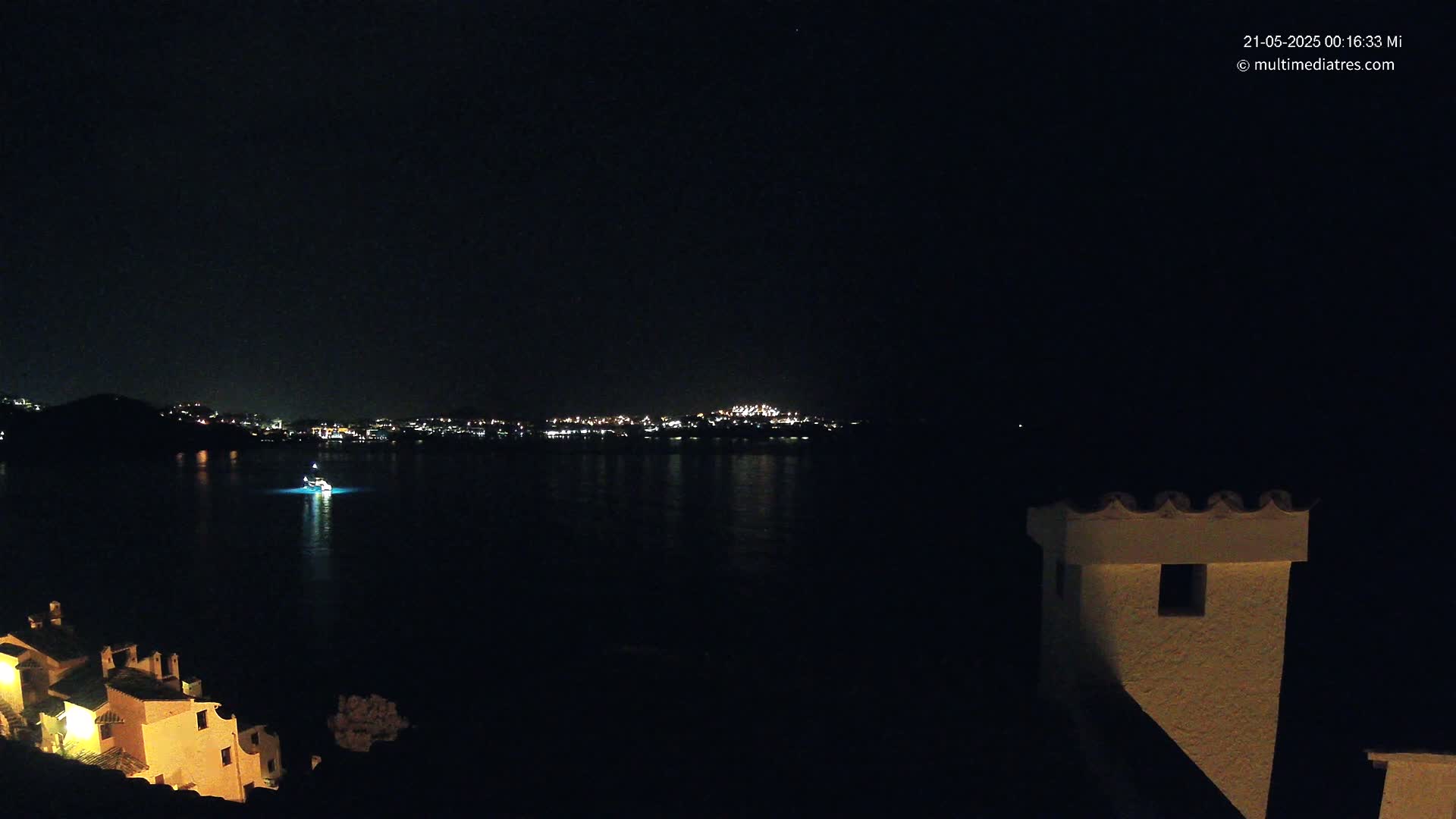 A nighttime view of a calm body of water with a distant illuminated cityscape and a small boat, partially obscured by darkness, seen from a vantage point near a building with illuminated windows.