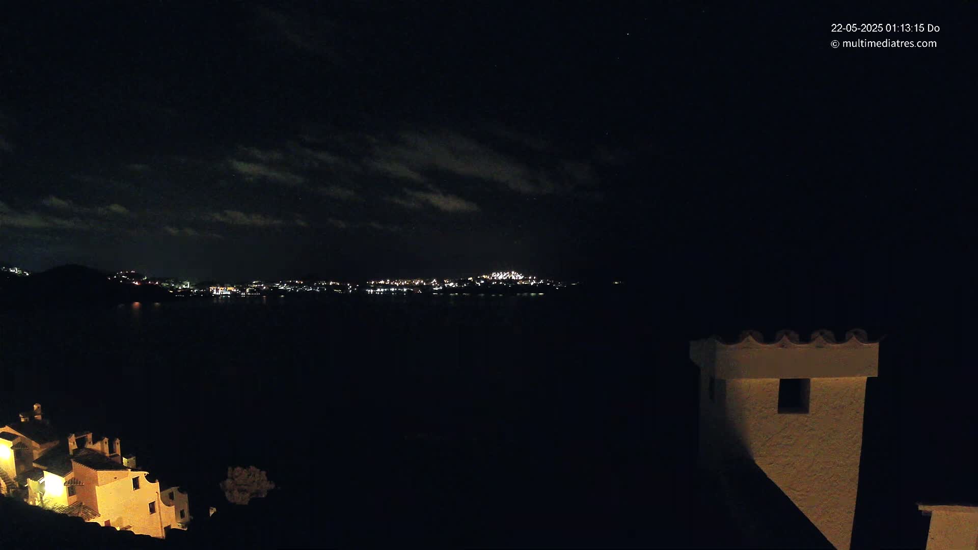 A nighttime view of a town's lights across a dark body of water, partially obscured by clouds, with a building in the foreground.