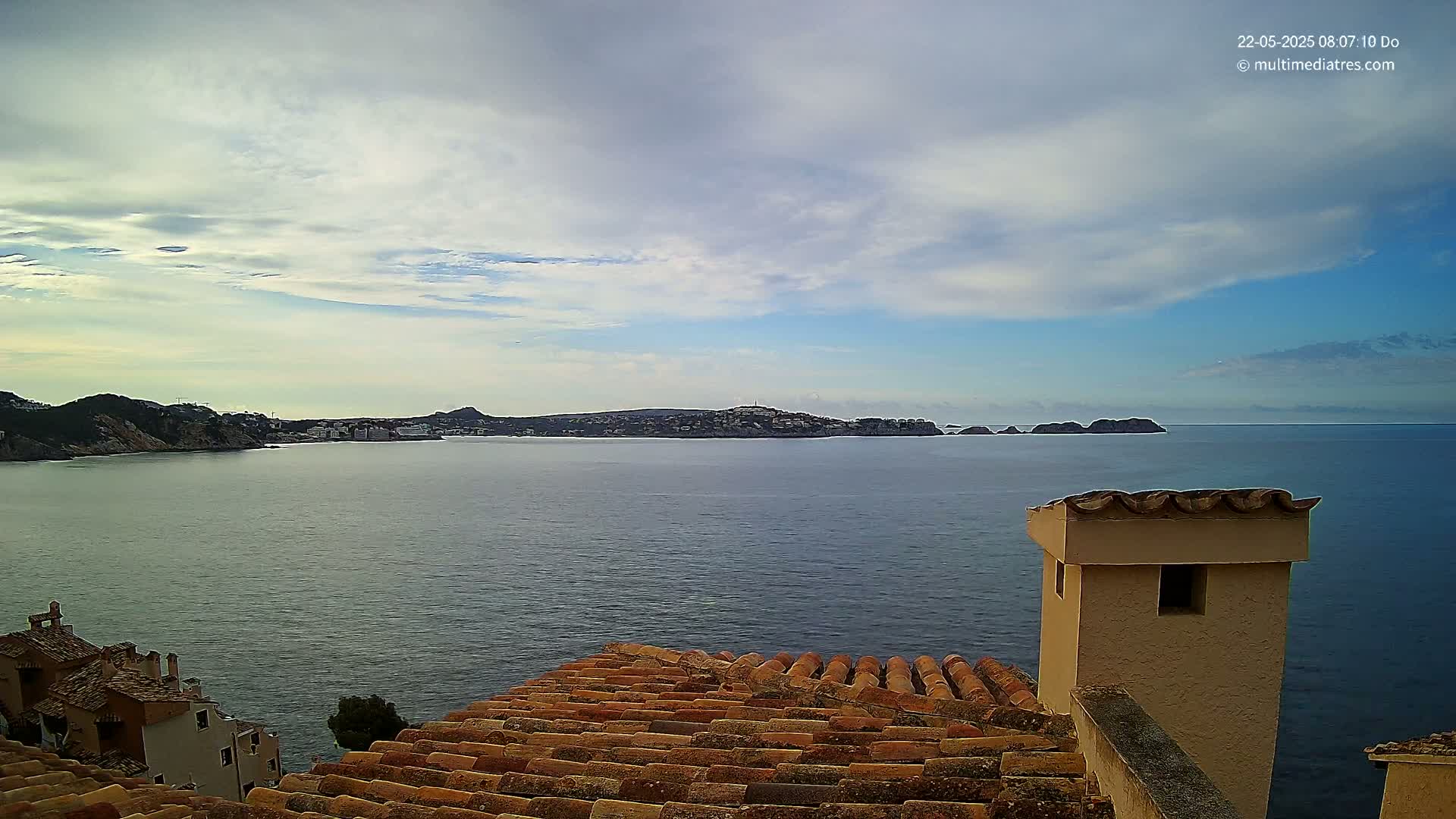 A partly cloudy sky overlooks a calm sea with a coastal town and islands in the distance, viewed from a terracotta rooftop with a chimney.