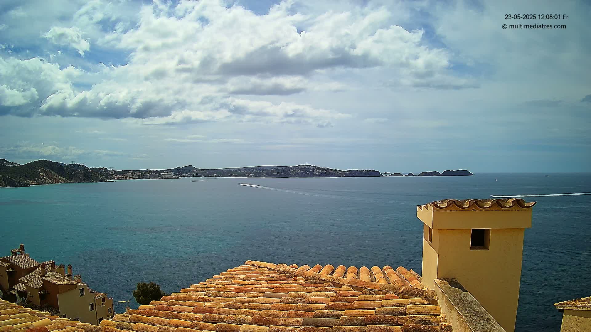 A partly cloudy sky overlooks a calm ocean bay from a terracotta roof with a tan chimney, showing a distant coastline and small boats.