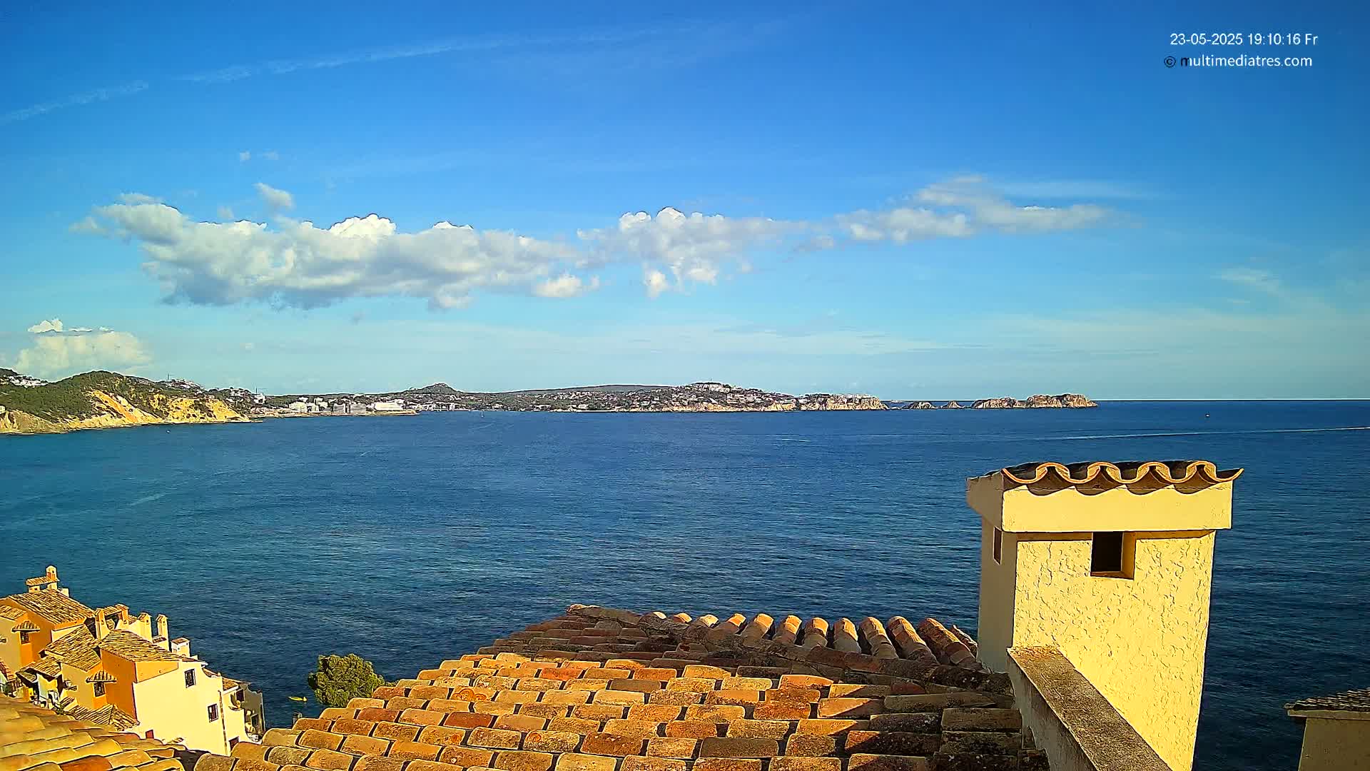 A partly cloudy blue sky overlooks a calm blue sea, with a coastal town and distant hills visible from a rooftop with terracotta tiles and a yellow chimney.
