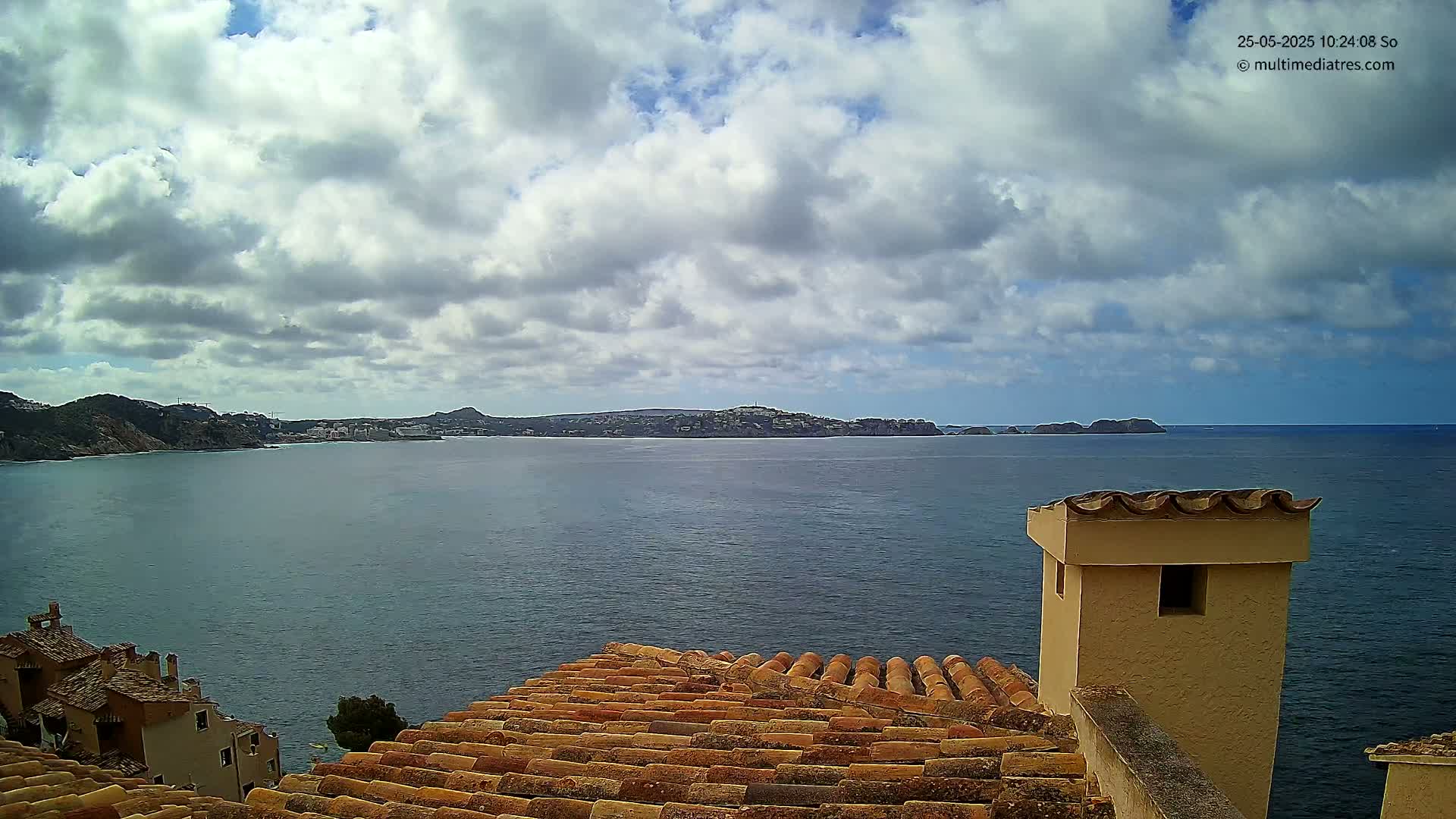 A partly cloudy sky overlooks a calm sea with distant hills and buildings, viewed from a tiled roof with a chimney.