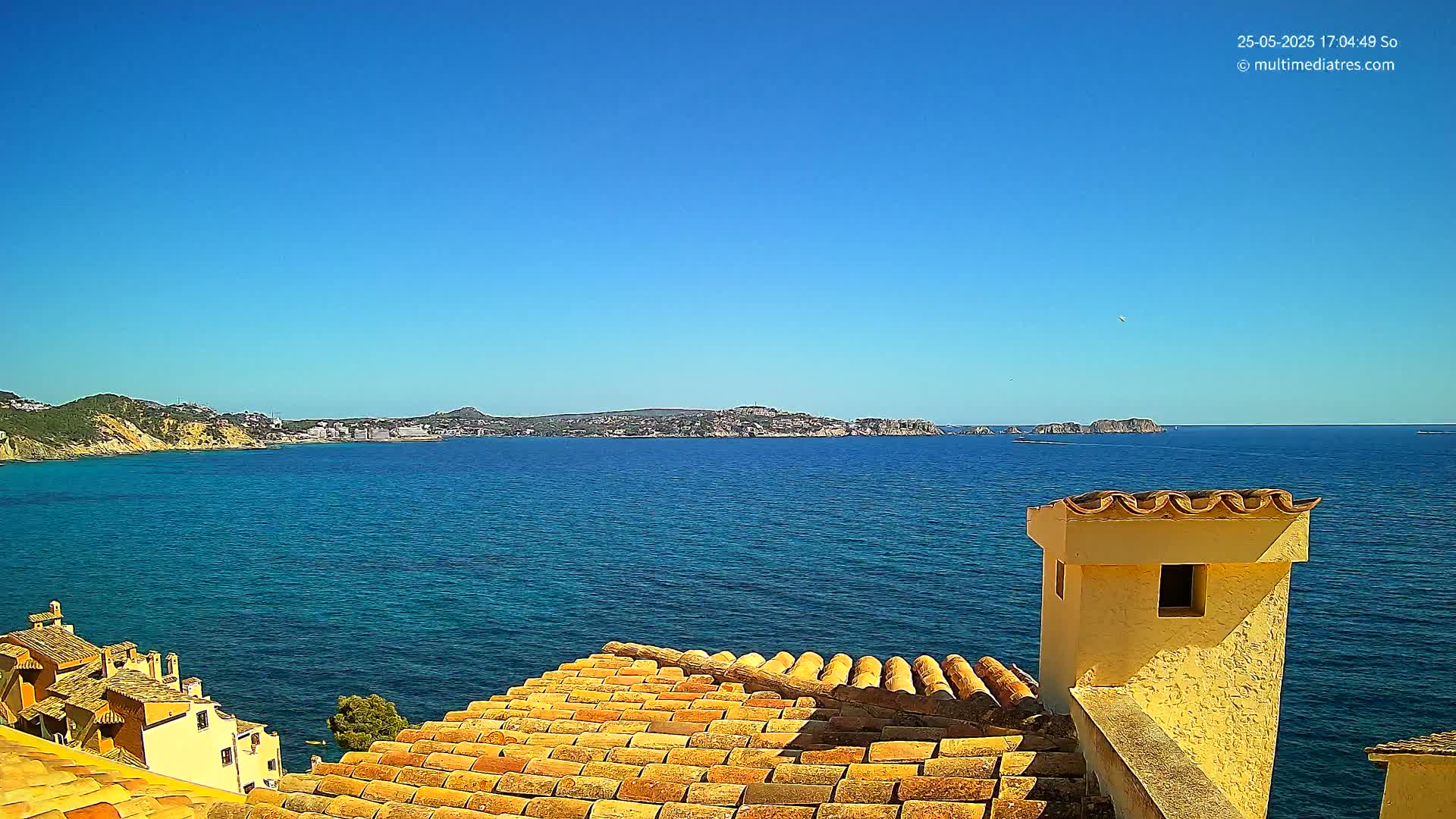 A sun-drenched coastal view reveals a terracotta roof overlooking a calm, azure sea and distant hills under a clear blue sky.
