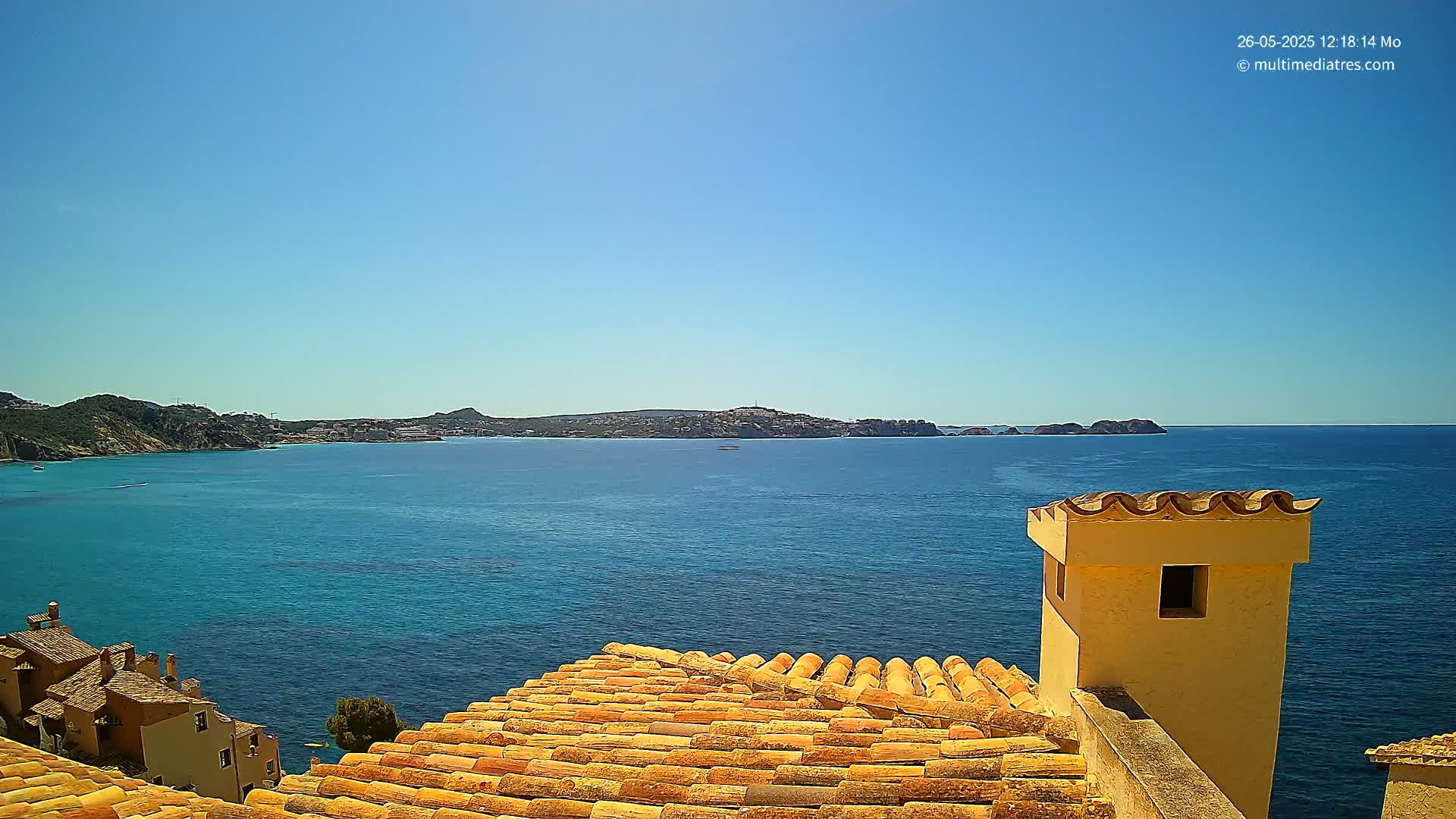 A terracotta roof overlooks a calm, blue ocean under a clear, sunny sky, with a distant coastline visible in the background.