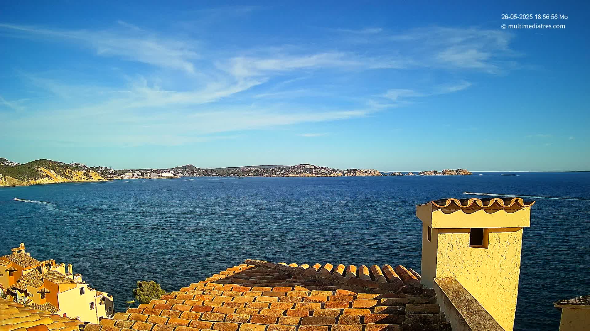 A view from a rooftop shows a calm blue sea under a mostly sunny sky, with a distant coastline and a few boats.
