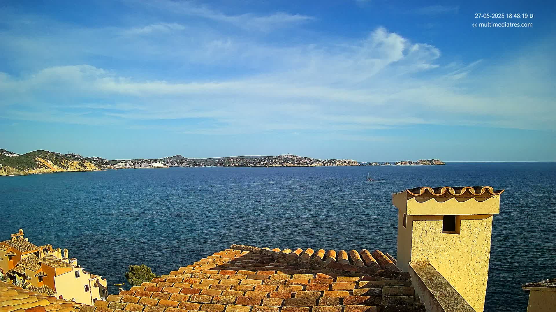 A view from a tiled roof shows a calm blue sea under a mostly sunny sky, with a distant coastline and buildings visible across the water.