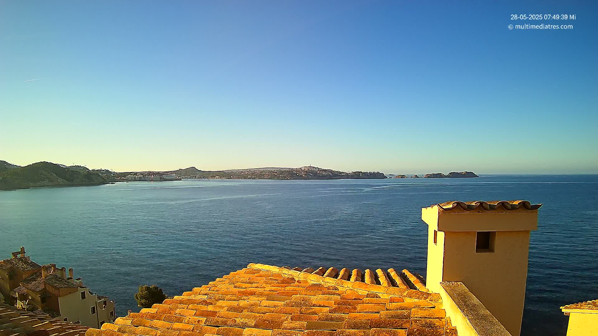 A clear blue sky overlooks a calm sea and distant land, viewed from a terracotta roof with a tan chimney.