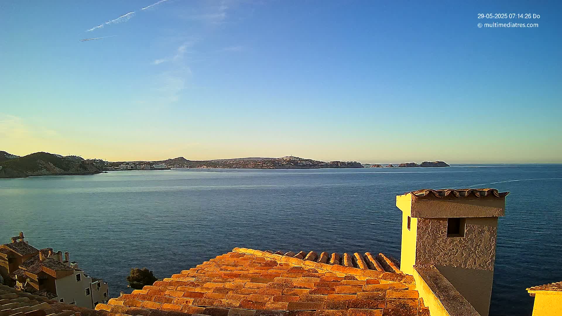A clear, calm sea is viewed from a terracotta roof, overlooking a distant town nestled between hills under a mostly sunny sky.