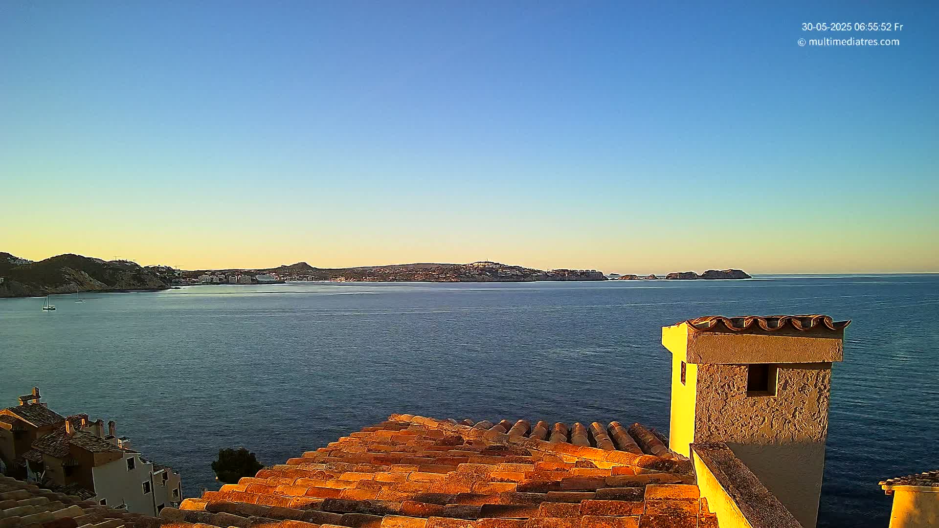 A coastal town's terracotta rooftops overlook a calm, blue sea under a clear, pale-blue sky at sunrise.