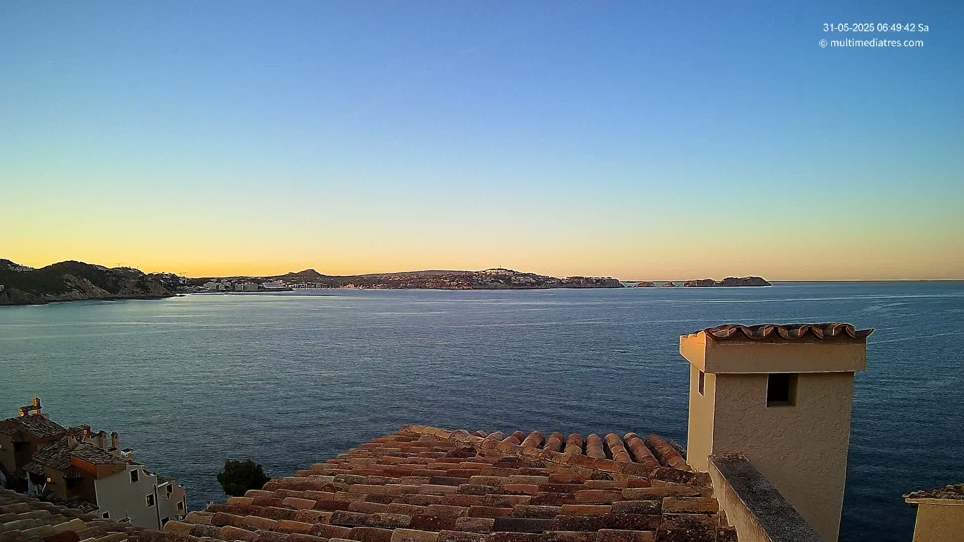A calm sea at sunset is viewed from a tiled rooftop, with a distant town nestled between hills visible on the horizon.