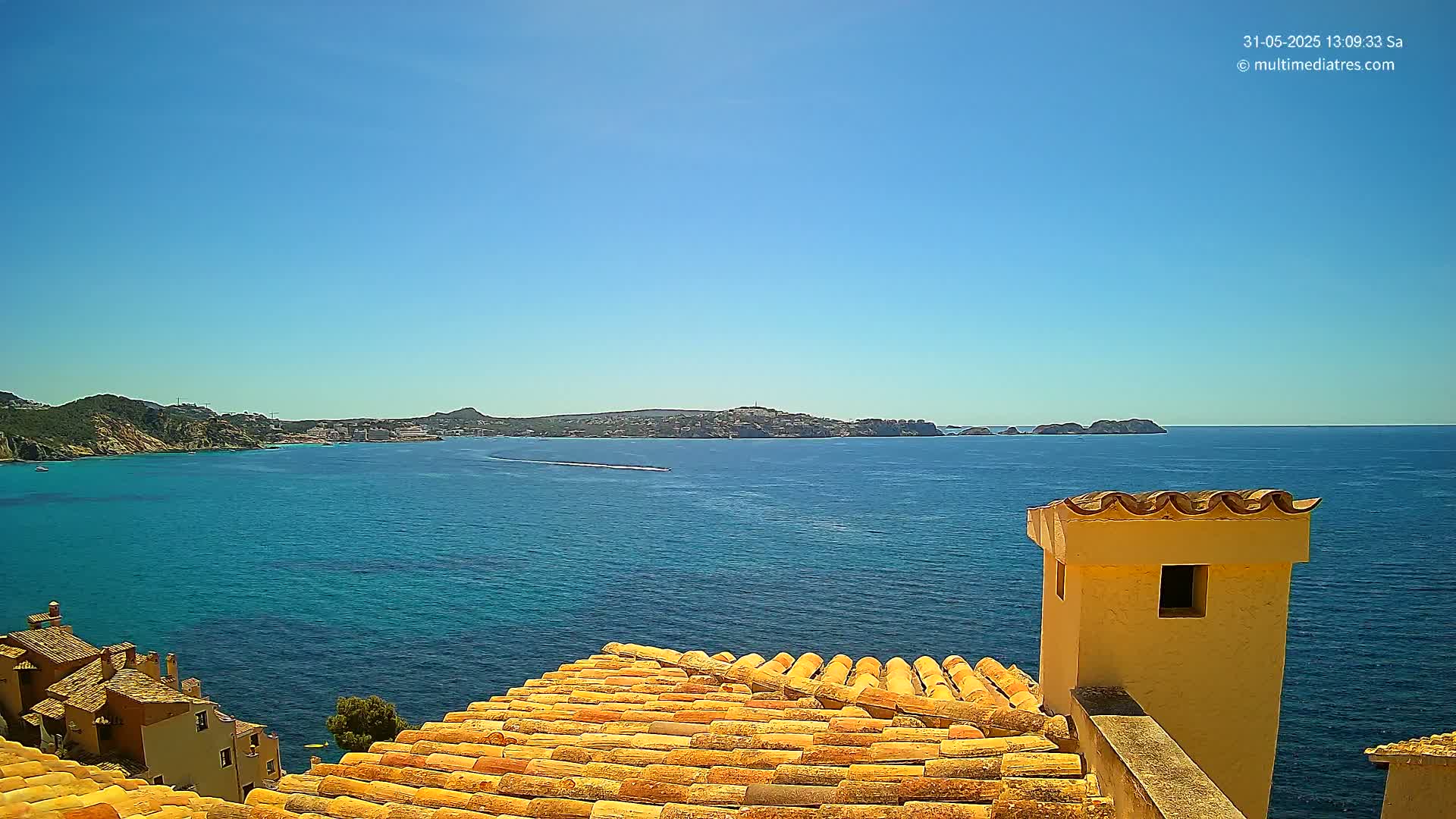 A clear, sunny day reveals a panoramic view of a turquoise ocean, a distant coastline with buildings, and a section of terracotta rooftops in the foreground.