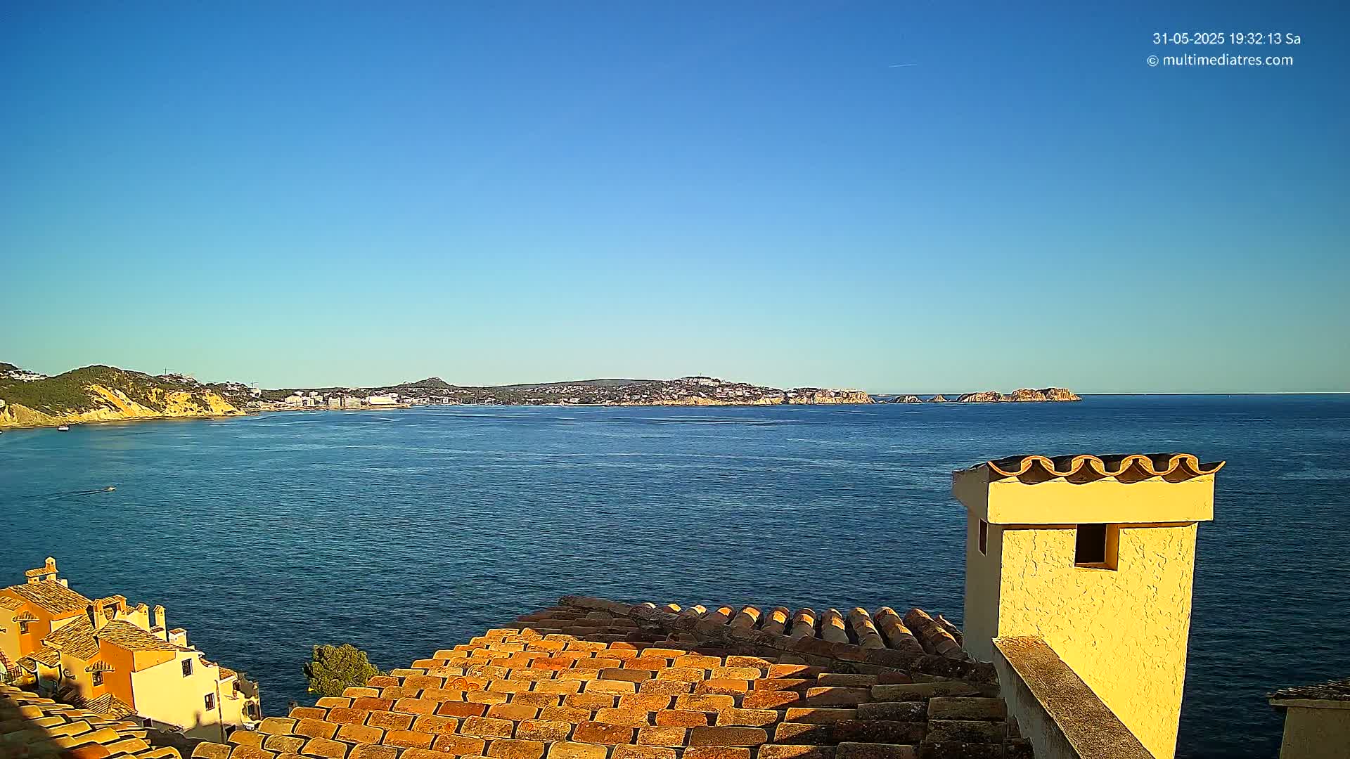A clear blue sky overlooks a calm blue sea separating a coastal town from a distant landmass with low hills, all viewed from a terracotta rooftop with a yellow chimney.