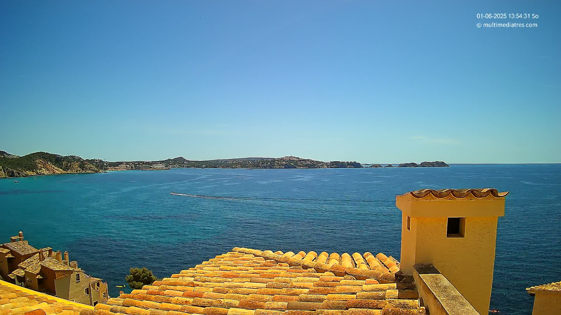 A clear blue sky overlooks a calm, turquoise sea with a distant coastline and islands, viewed from a rooftop with terracotta tiles and a pale yellow chimney.