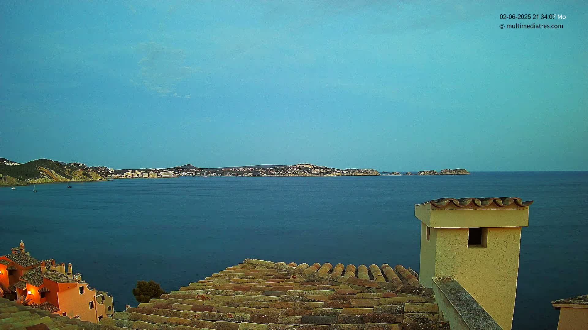 A tranquil coastal scene at dusk shows a calm blue sea with a distant town and islands under a clear sky, viewed from a rooftop with terracotta tiles and a light-yellow chimney.