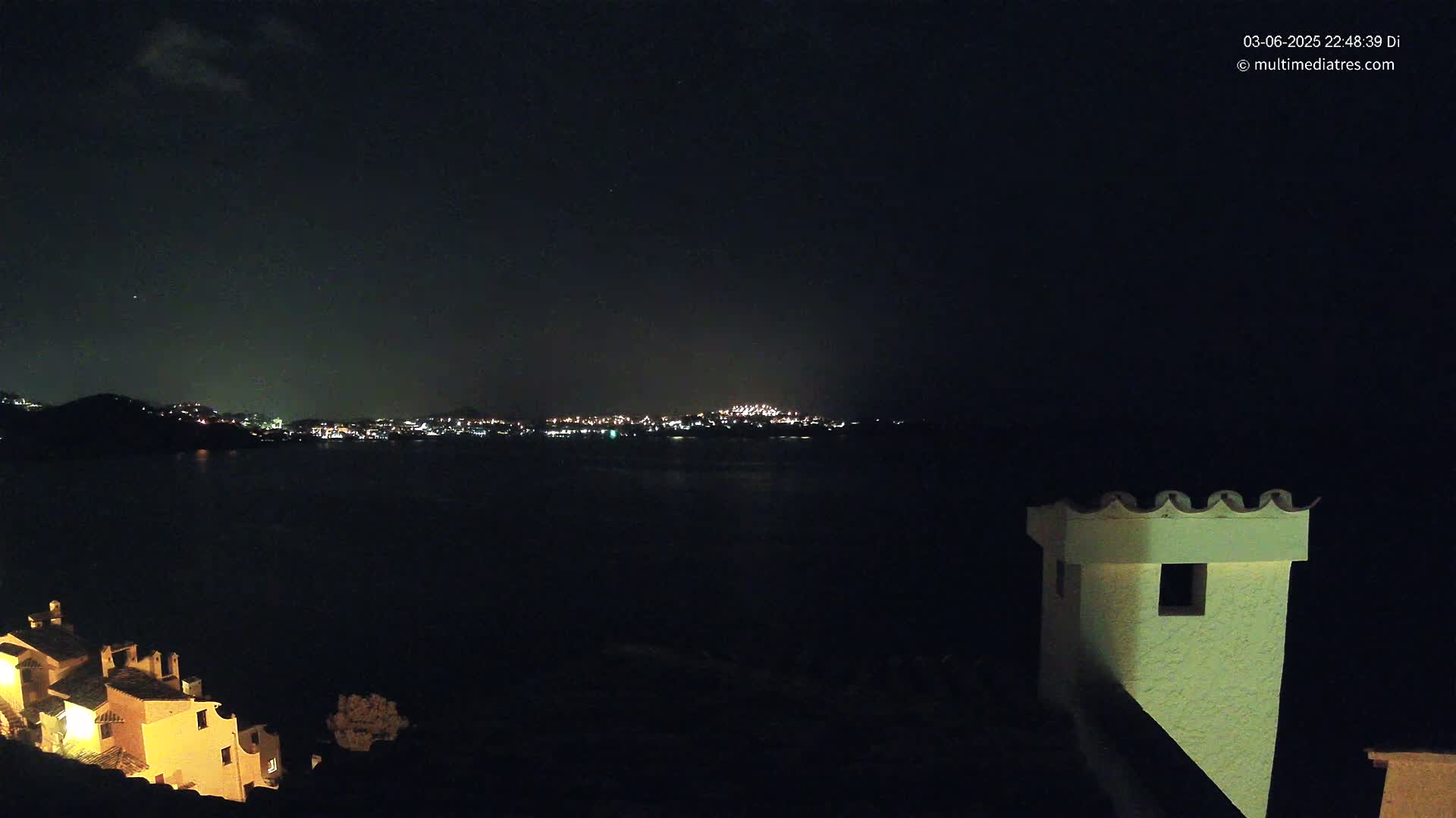 A nighttime view of a dark body of water with a distant city skyline illuminated by lights, partially obscured by a dark hill, with a portion of a building and its chimney in the foreground under a mostly clear sky.