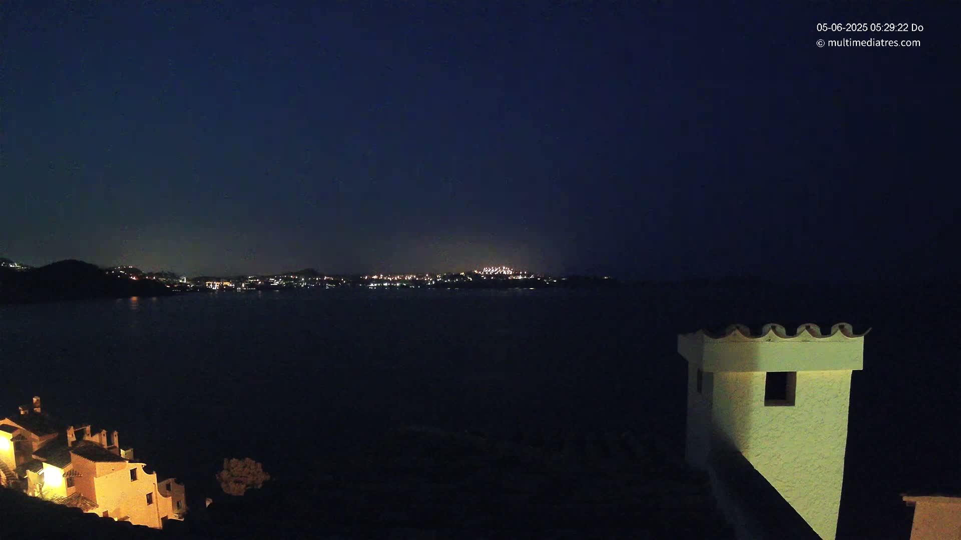 A nighttime coastal scene shows a dark body of water separating a small, lit village from a larger, more distant city across the water under a clear, dark sky.