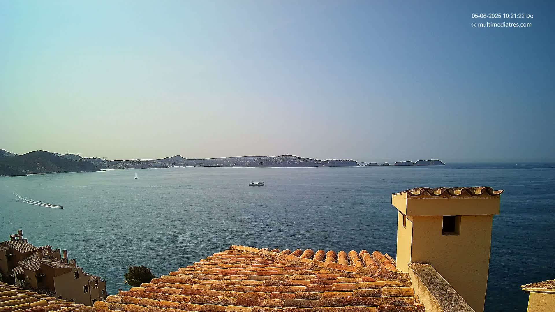 A terracotta roof overlooks a calm blue sea with distant hills and a few small boats under a clear sky.