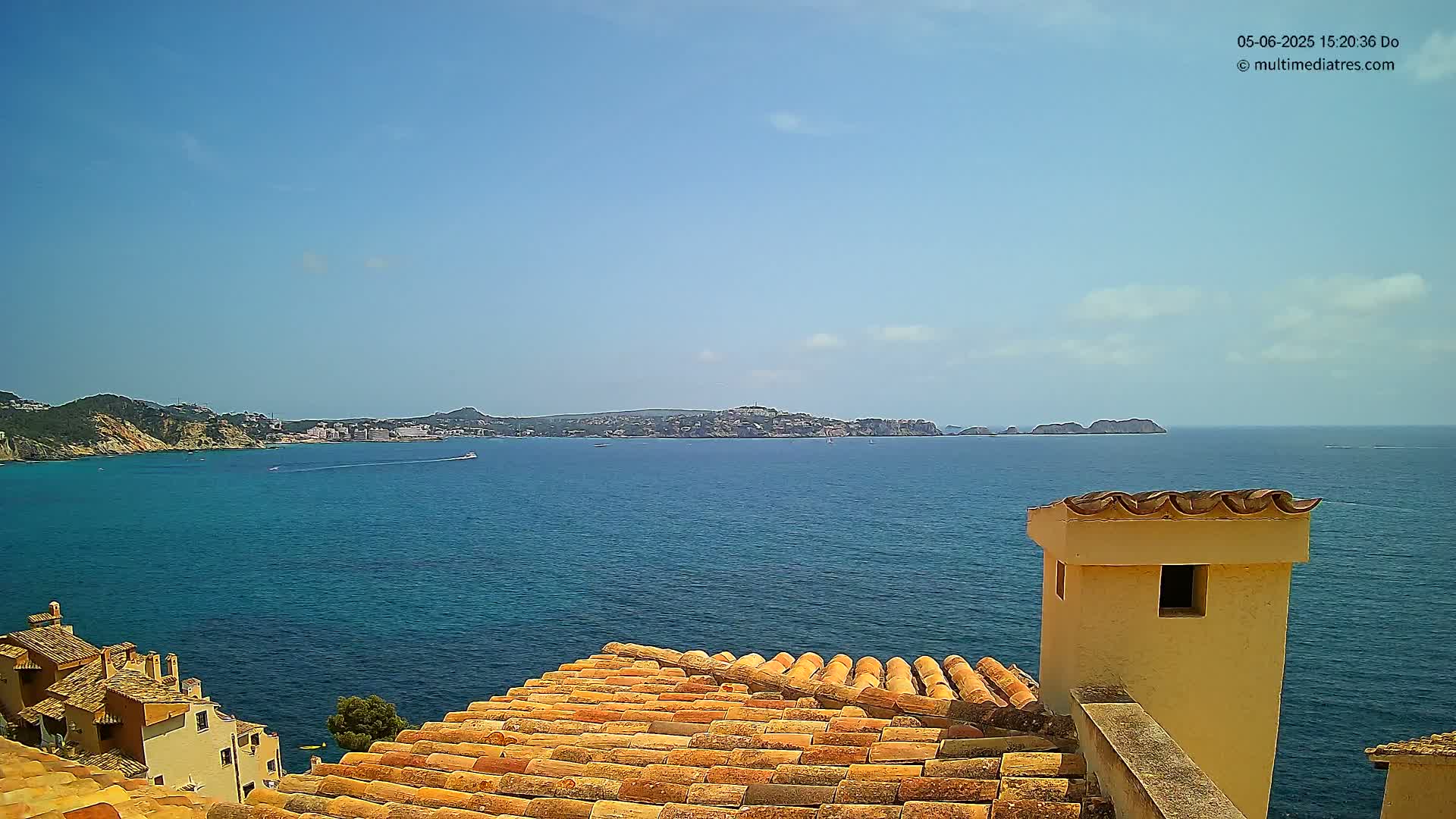 A terracotta roof overlooks a calm, blue ocean with a distant coastline under a mostly clear, sunny sky.