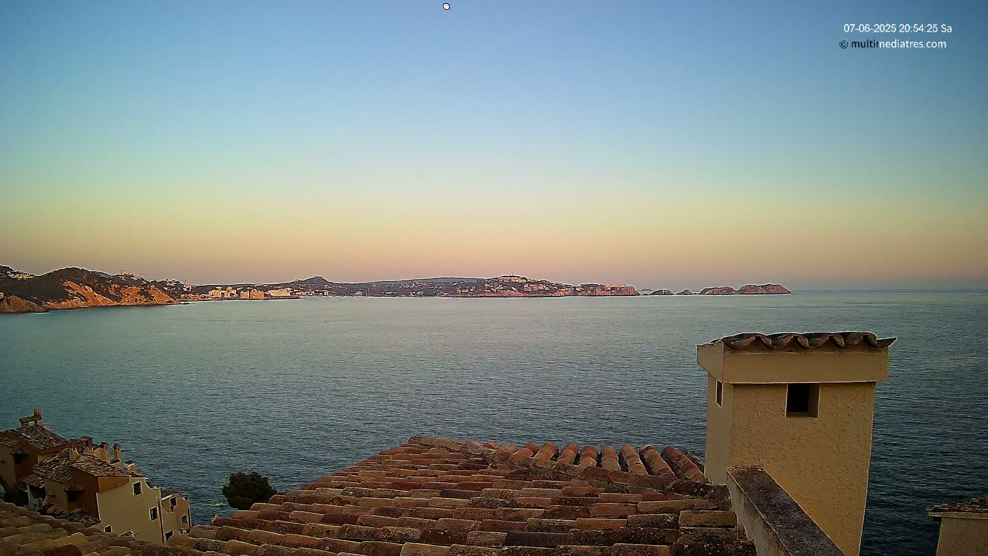 A tranquil coastal scene at sunset shows a calm sea with distant hills and a portion of a tiled roof and chimney in the foreground.