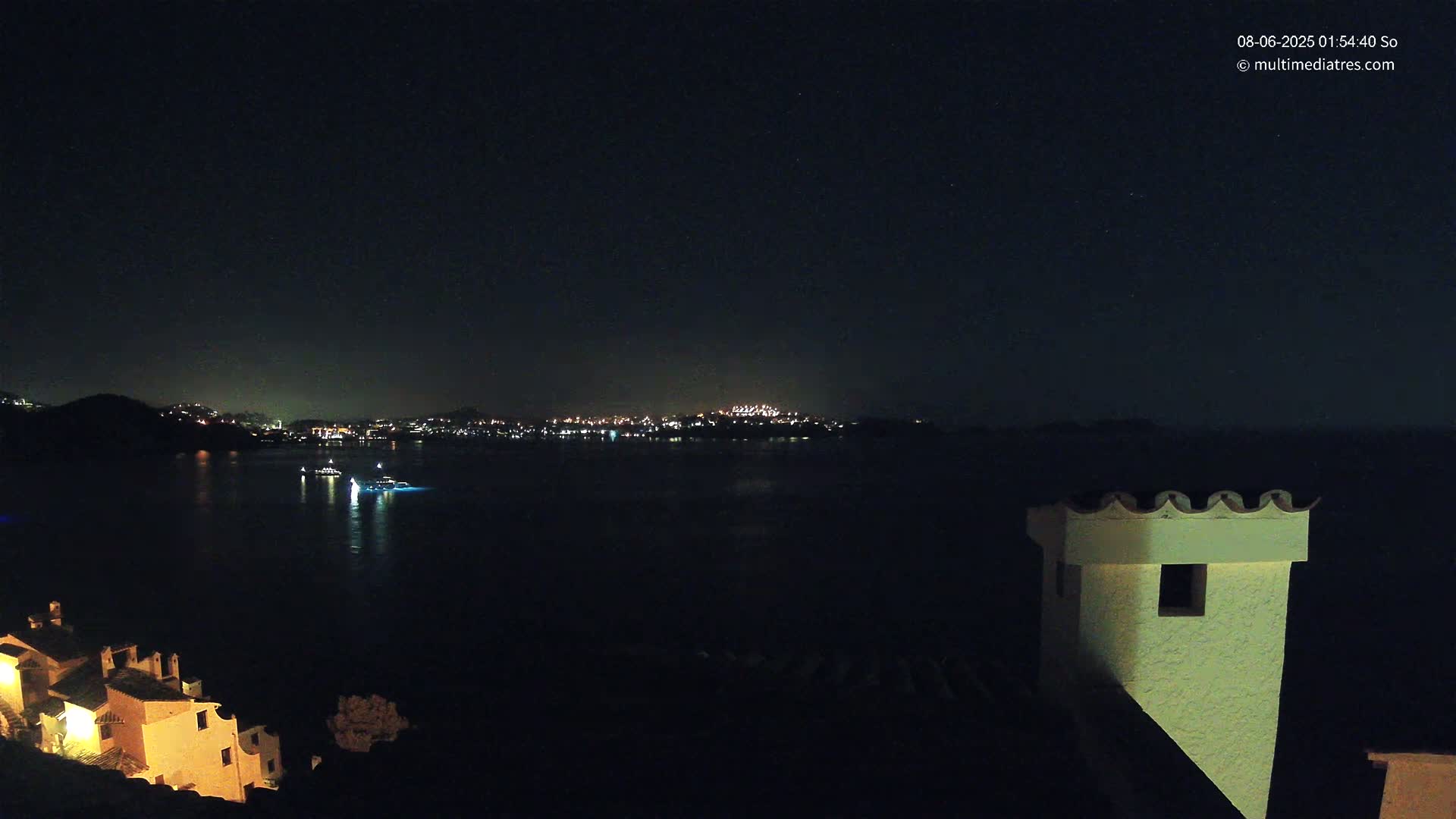 A nighttime view of a dark body of water with a distant city skyline illuminated, a few boats on the water, and a portion of a building in the foreground under a clear, starry sky.