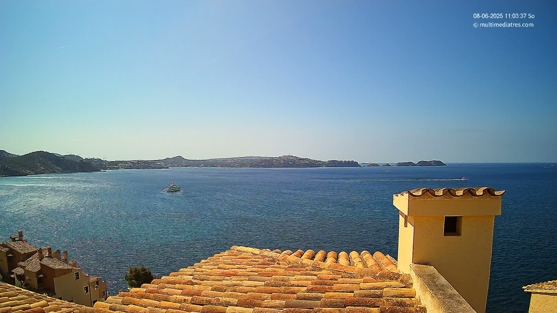 A terracotta roof overlooks a calm blue sea and distant coastline under a clear, sunny sky.