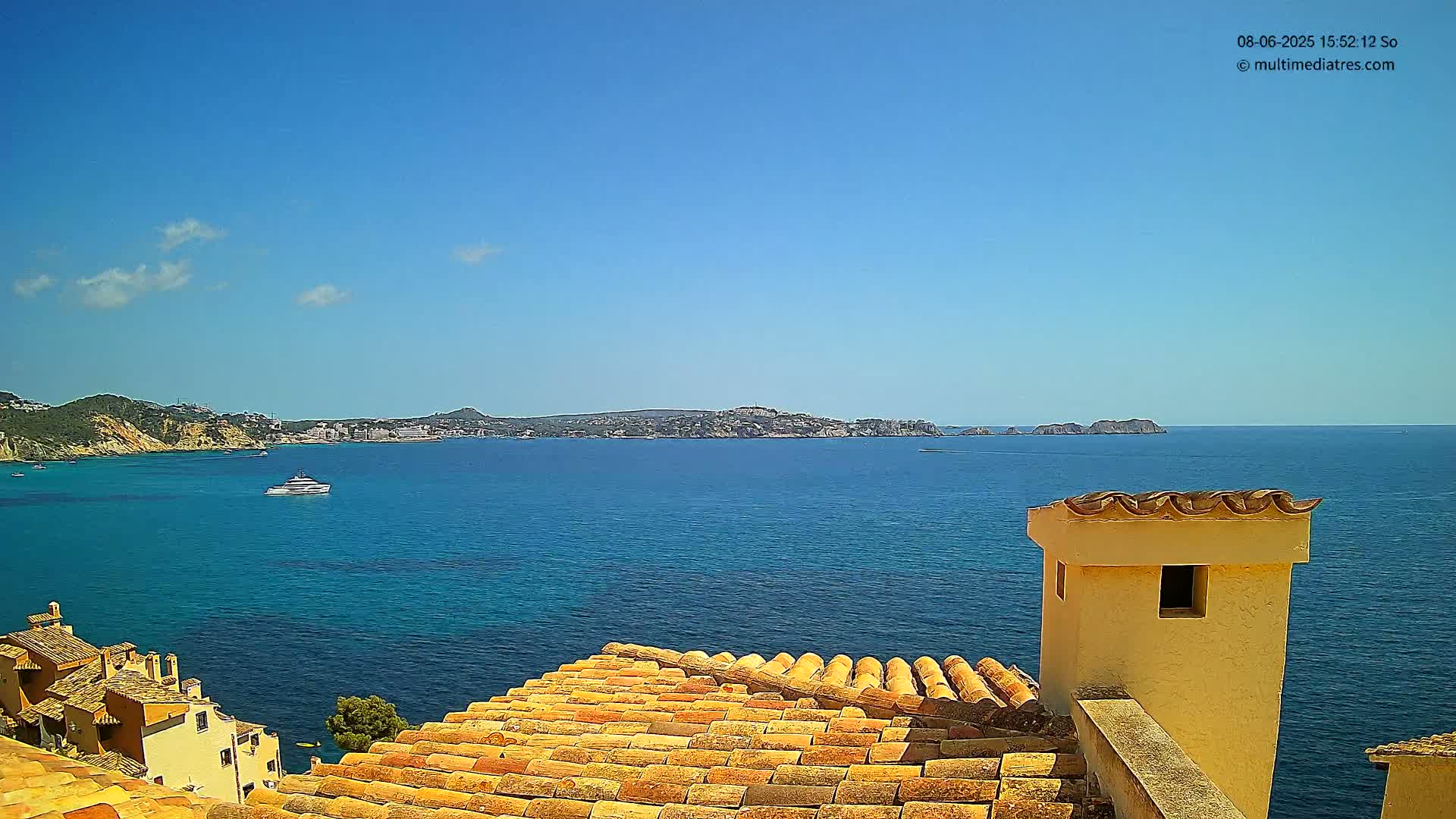 A terracotta roof overlooks a calm, blue ocean on a sunny day, with distant hills and a boat visible on the water.