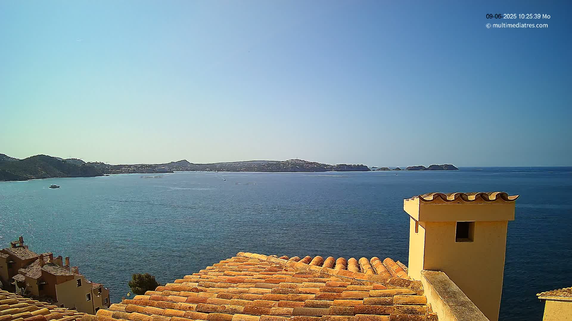 A terracotta roof overlooks a calm blue sea with distant hills and a clear sky.