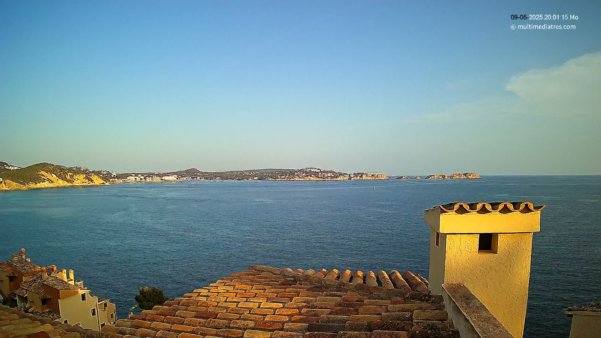 A calm, clear blue sea stretches out from a terracotta roof toward a distant coastline under a mostly clear sky.