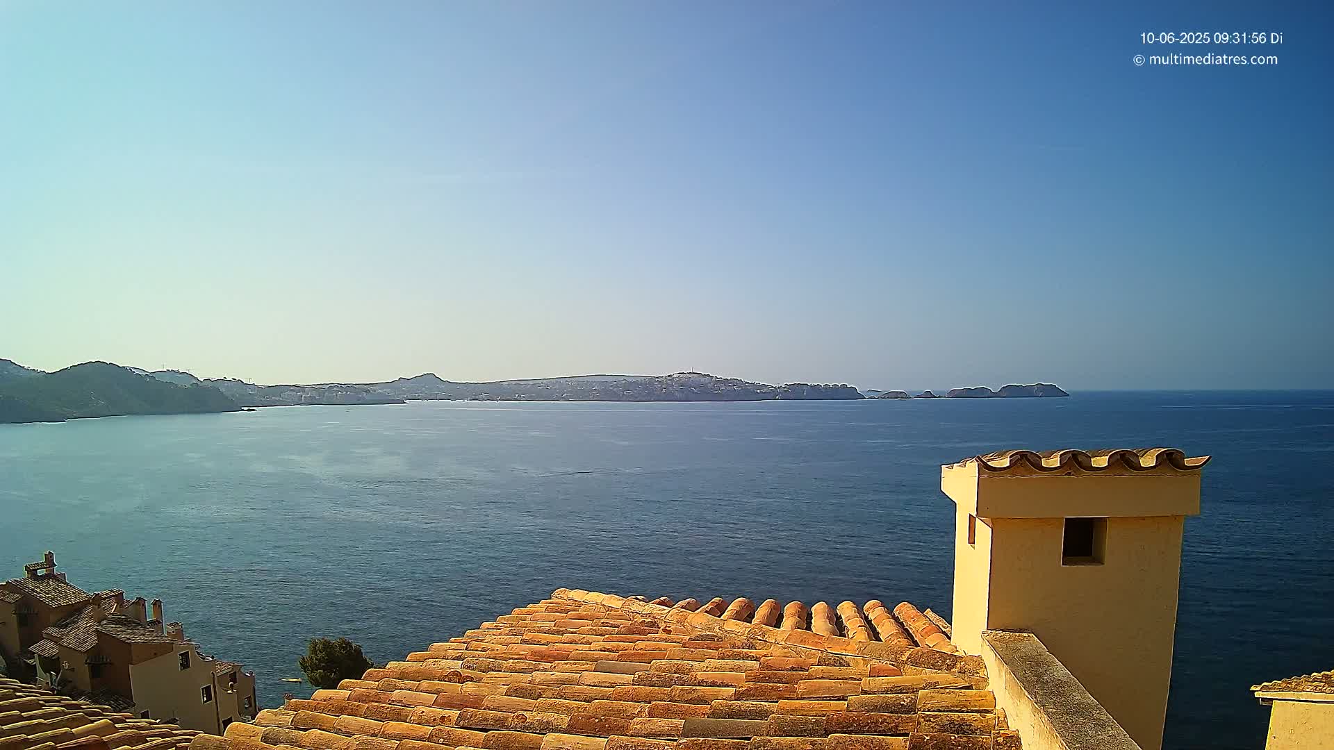 A terracotta roof overlooks a calm, blue sea with distant hills and islands under a clear sky.