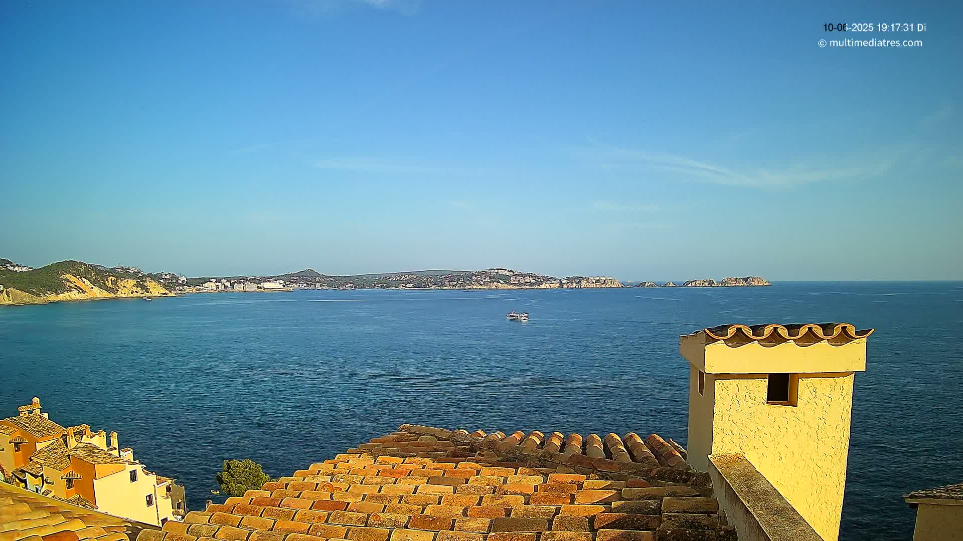 A terracotta roof overlooks a calm blue sea, with a distant coastline and clear blue sky.