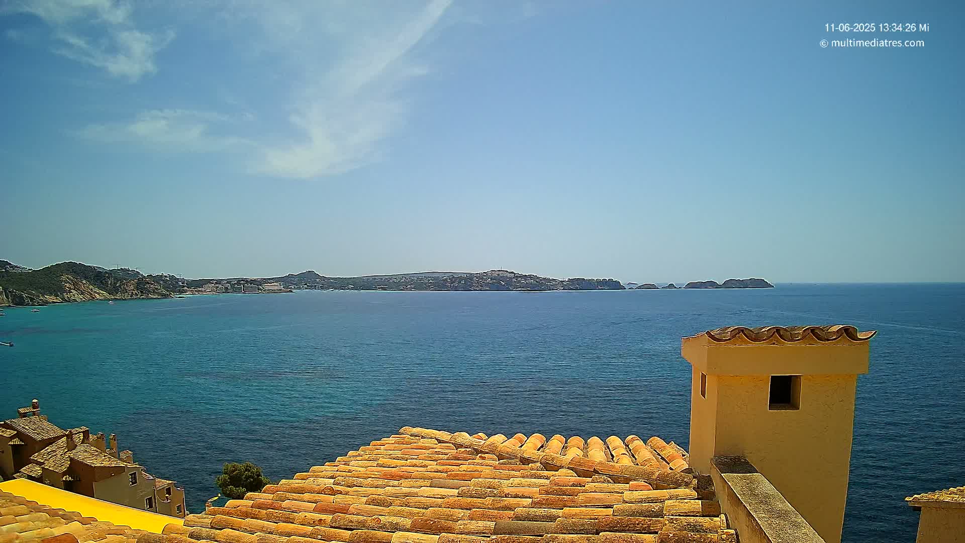 A terracotta roof overlooks a calm, blue sea with a distant coastline under a mostly sunny sky.
