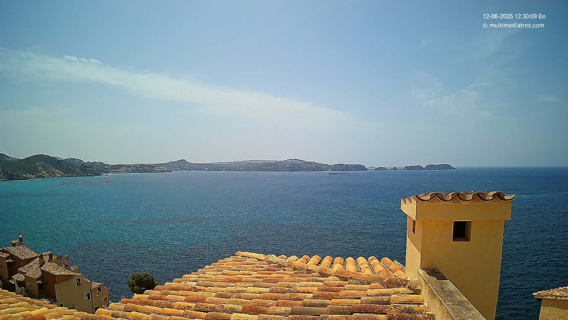 A terracotta roof overlooks a calm, blue ocean under a mostly clear, sunny sky, with distant hills and islands visible on the horizon.