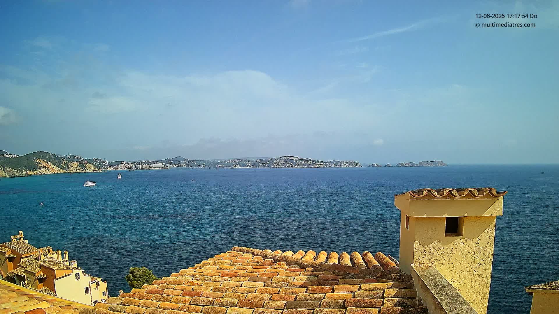 A tiled roof overlooks a calm blue sea on a sunny day, with distant landmasses and a few boats visible.