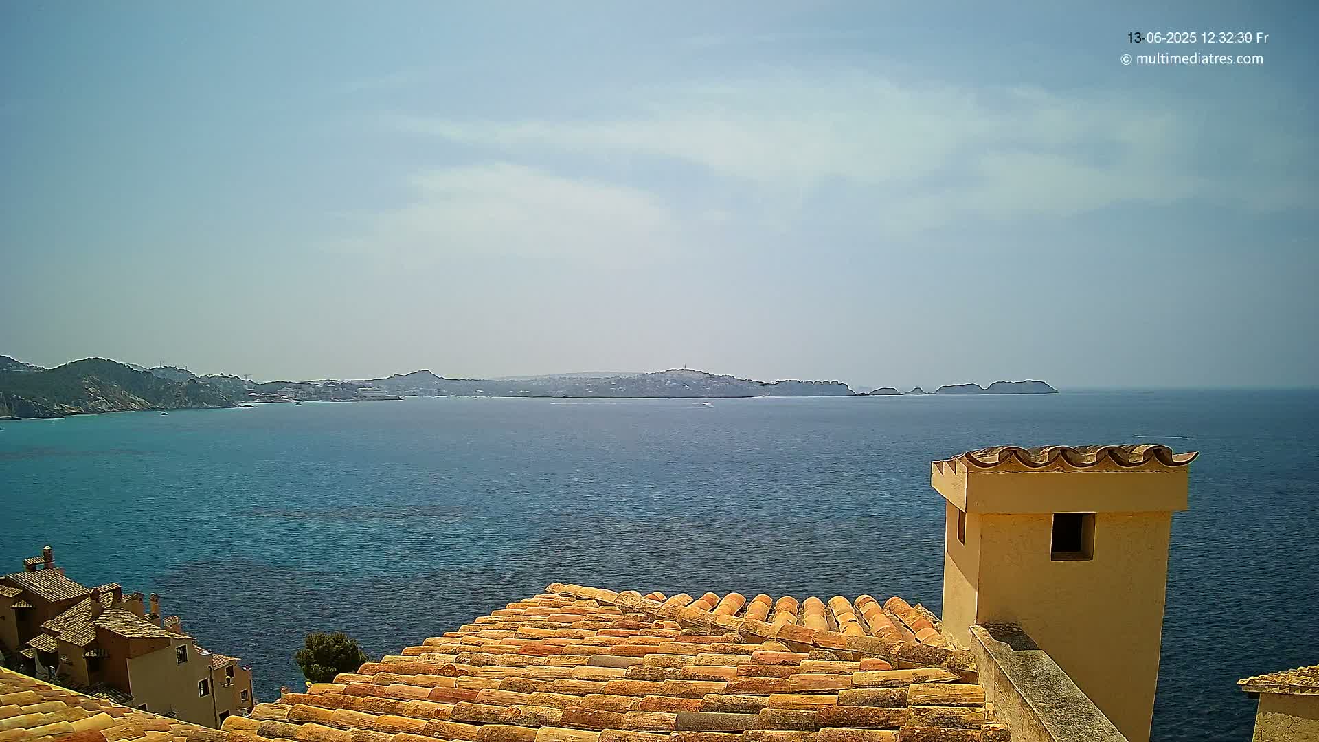 A terracotta roof overlooks a calm, blue sea with distant landmasses under a mostly clear sky.