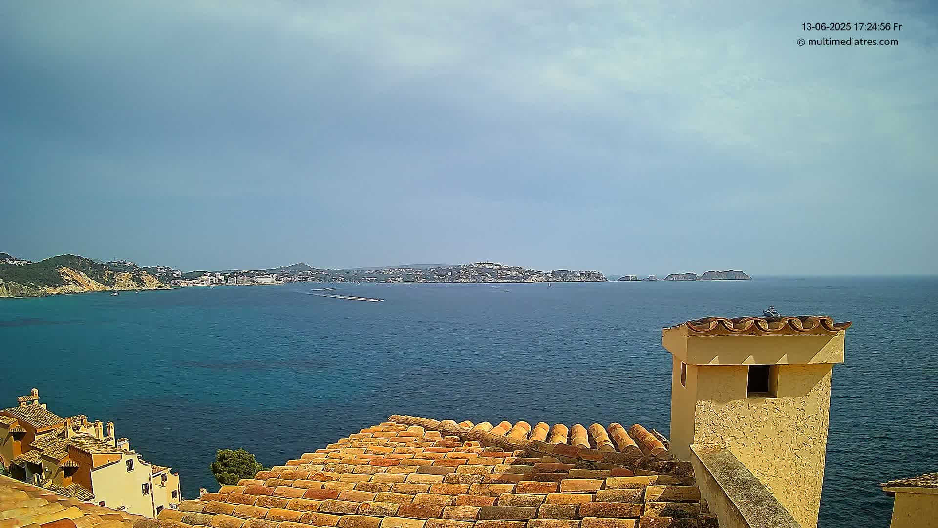 A partly cloudy day reveals a coastal view of a town with terracotta rooftops overlooking a calm blue sea and distant islands.