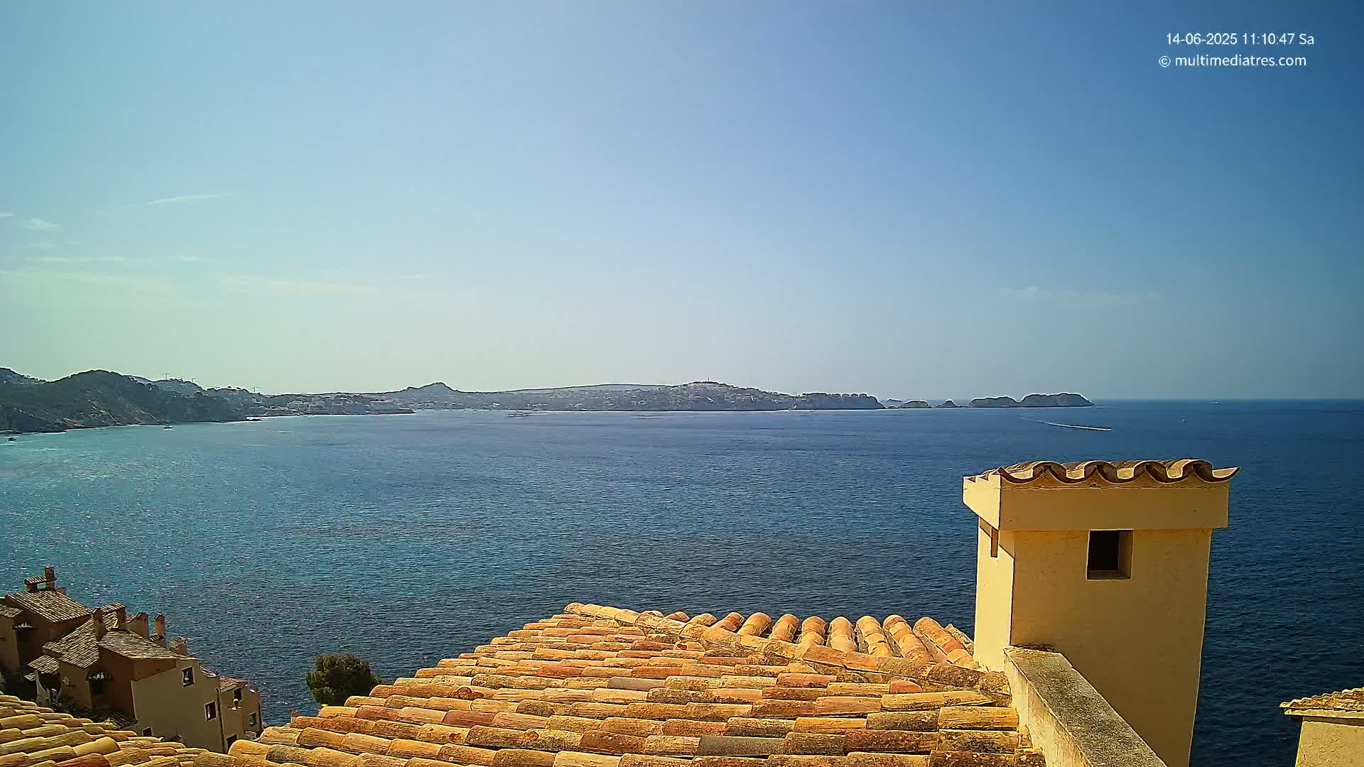 A terracotta roof and a pale yellow chimney overlook a calm, blue sea with a distant coastline under a clear, sunny sky.