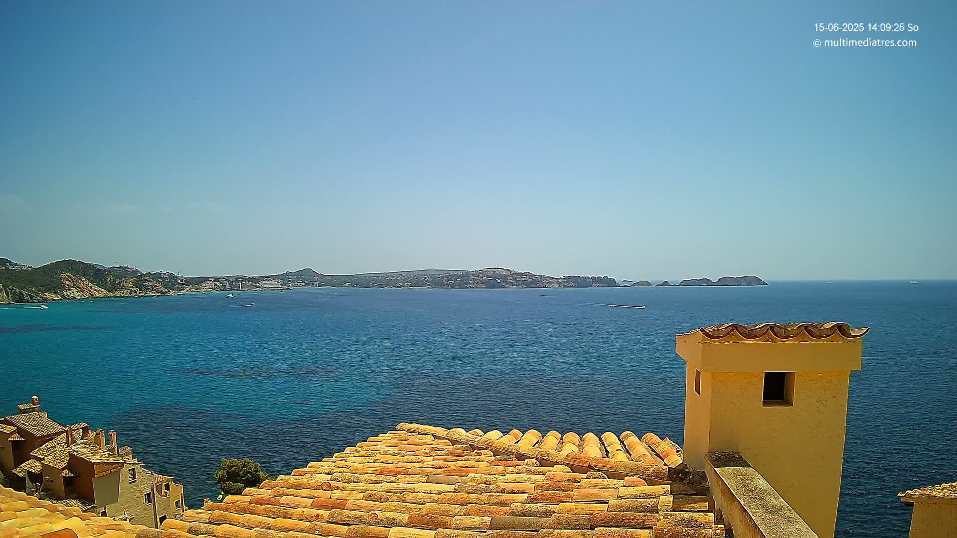 A clear blue sky overlooks a calm, deep blue sea, with a coastal town and distant islands visible from an orange tiled rooftop with a light-yellow chimney.