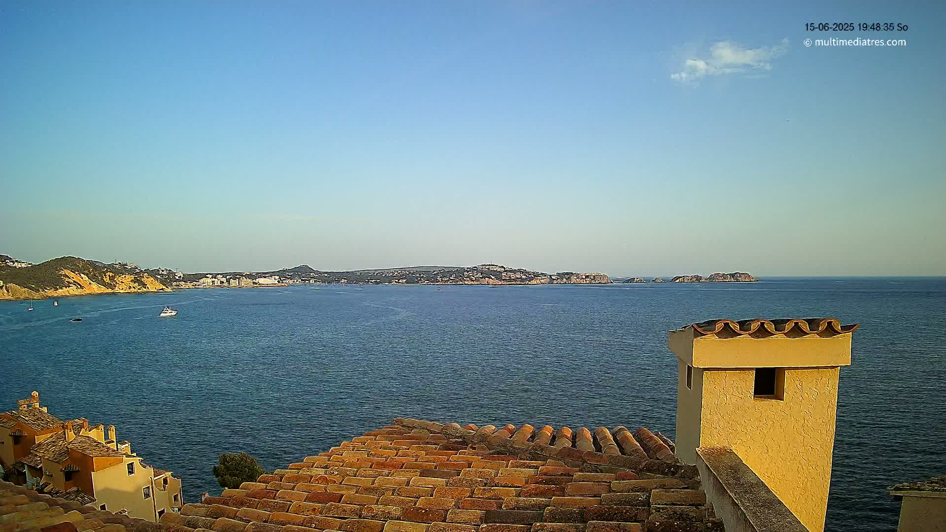 A tranquil seaside view under a clear blue sky shows a calm body of water, distant islands, and a section of terracotta rooftops with a chimney in the foreground.