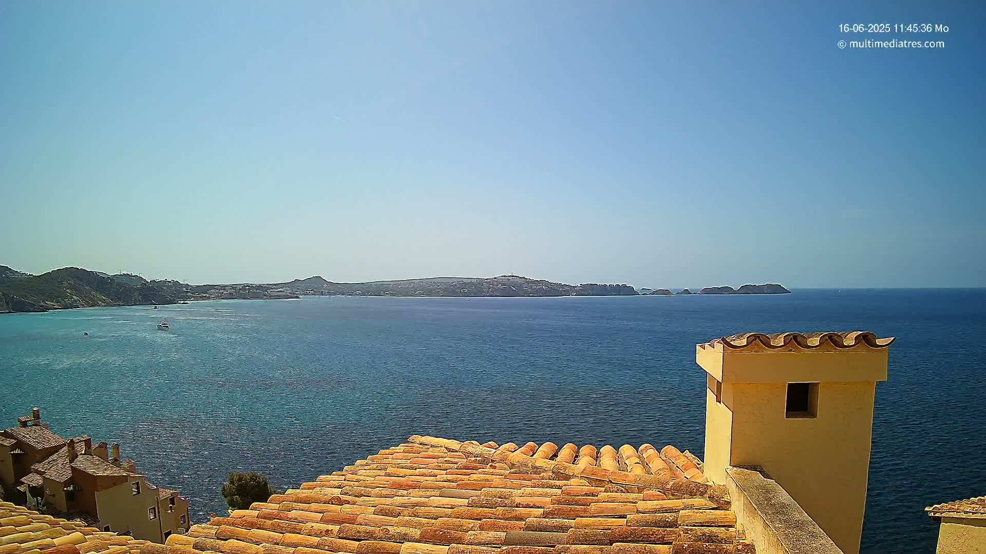 A tiled roof overlooks a calm blue sea and distant coastline under a clear, sunny sky.
