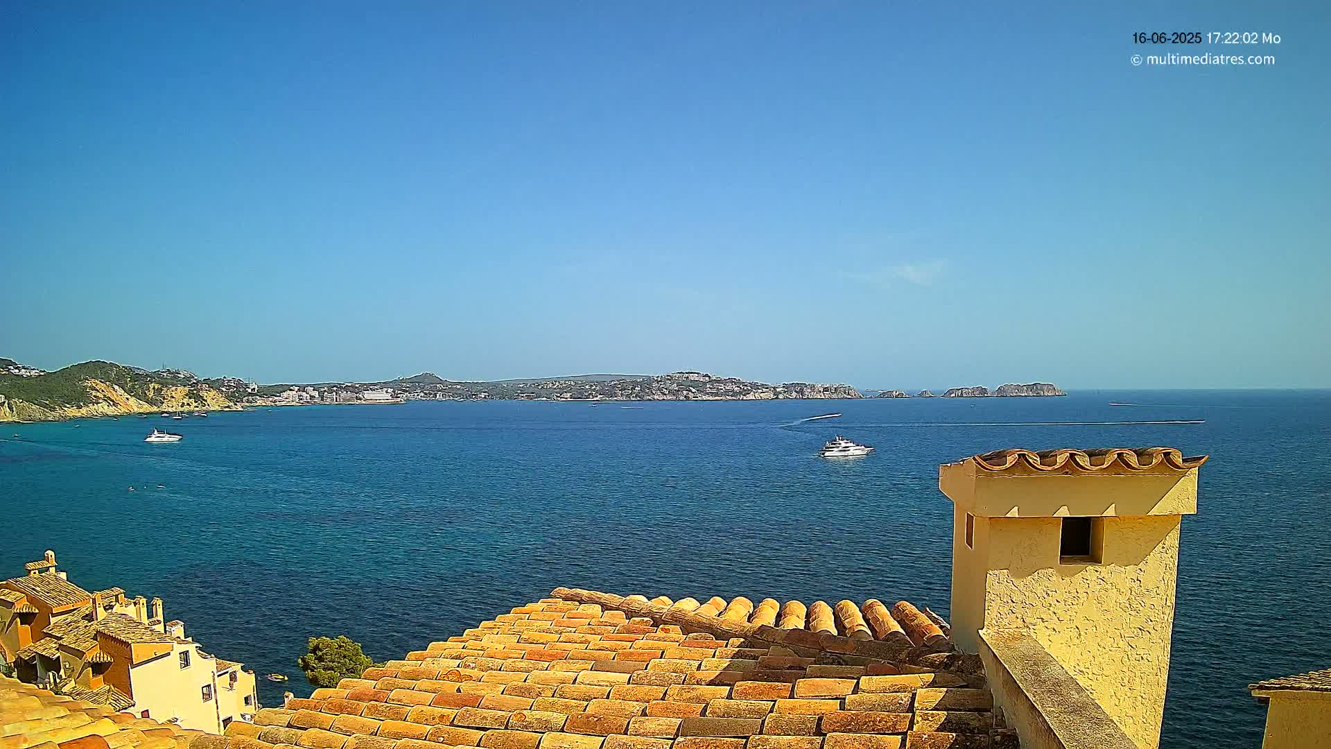 A clear blue sky overlooks a calm blue sea with a few boats, a distant coastline, and a portion of a terracotta-tiled roof with a yellow chimney in the foreground.
