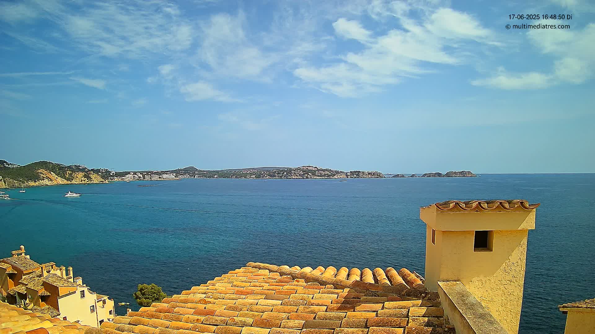 A terracotta roof overlooks a calm, blue sea with a distant coastline under a mostly sunny sky.