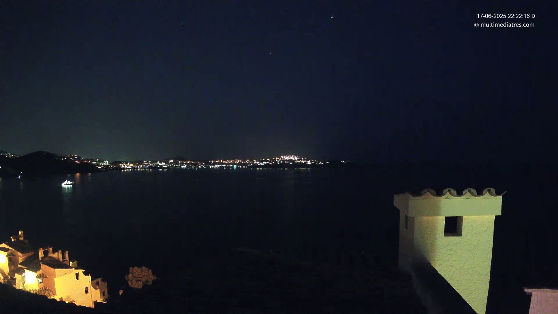 A nighttime view of a city's lights across a dark body of water, partially obscured by a hill, with a building and its chimney in the foreground under a clear, starry sky.