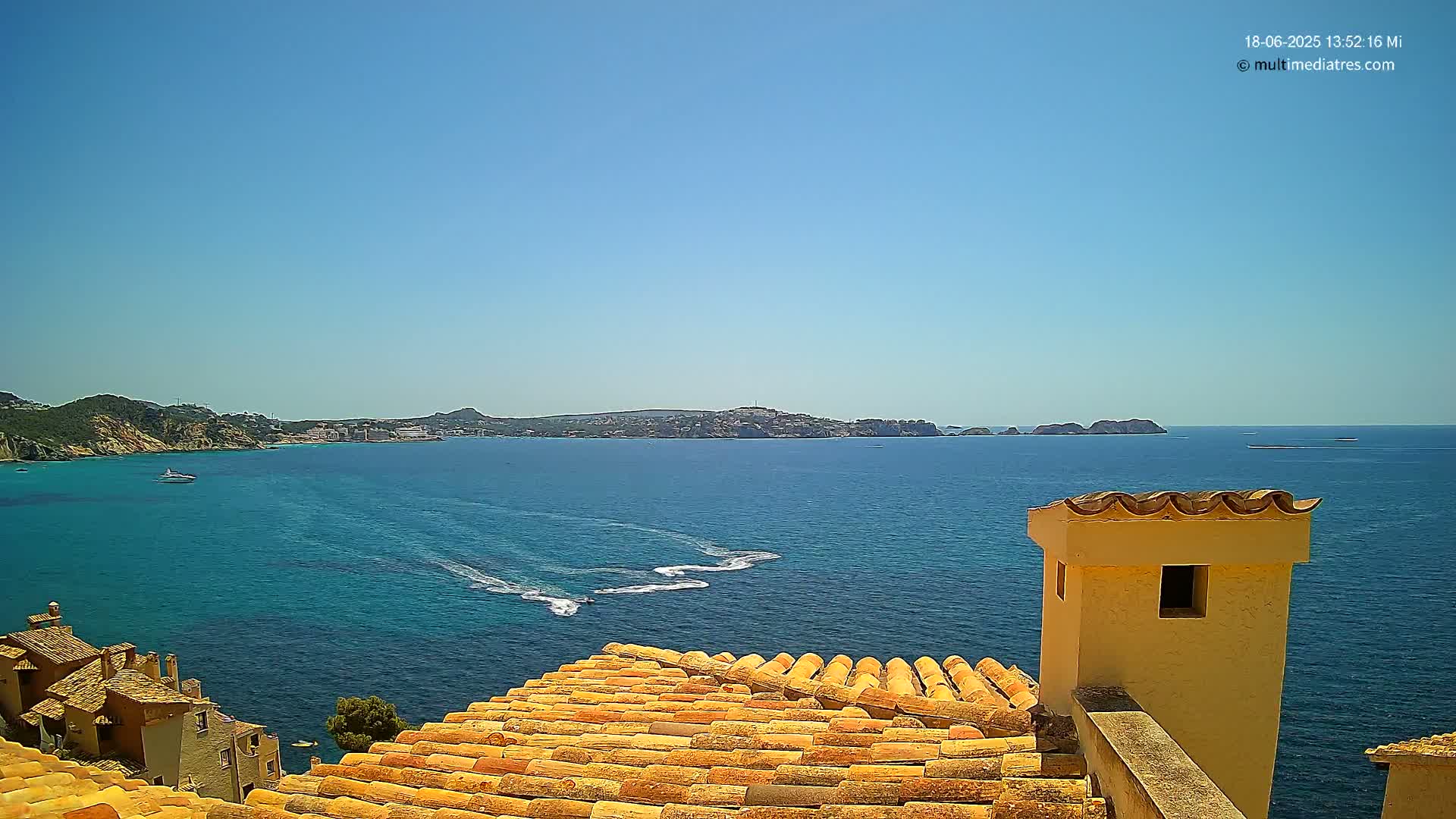 A sunny, seaside view from a rooftop shows a calm, blue sea with boats, a distant coastline, and terracotta roof tiles in the foreground.