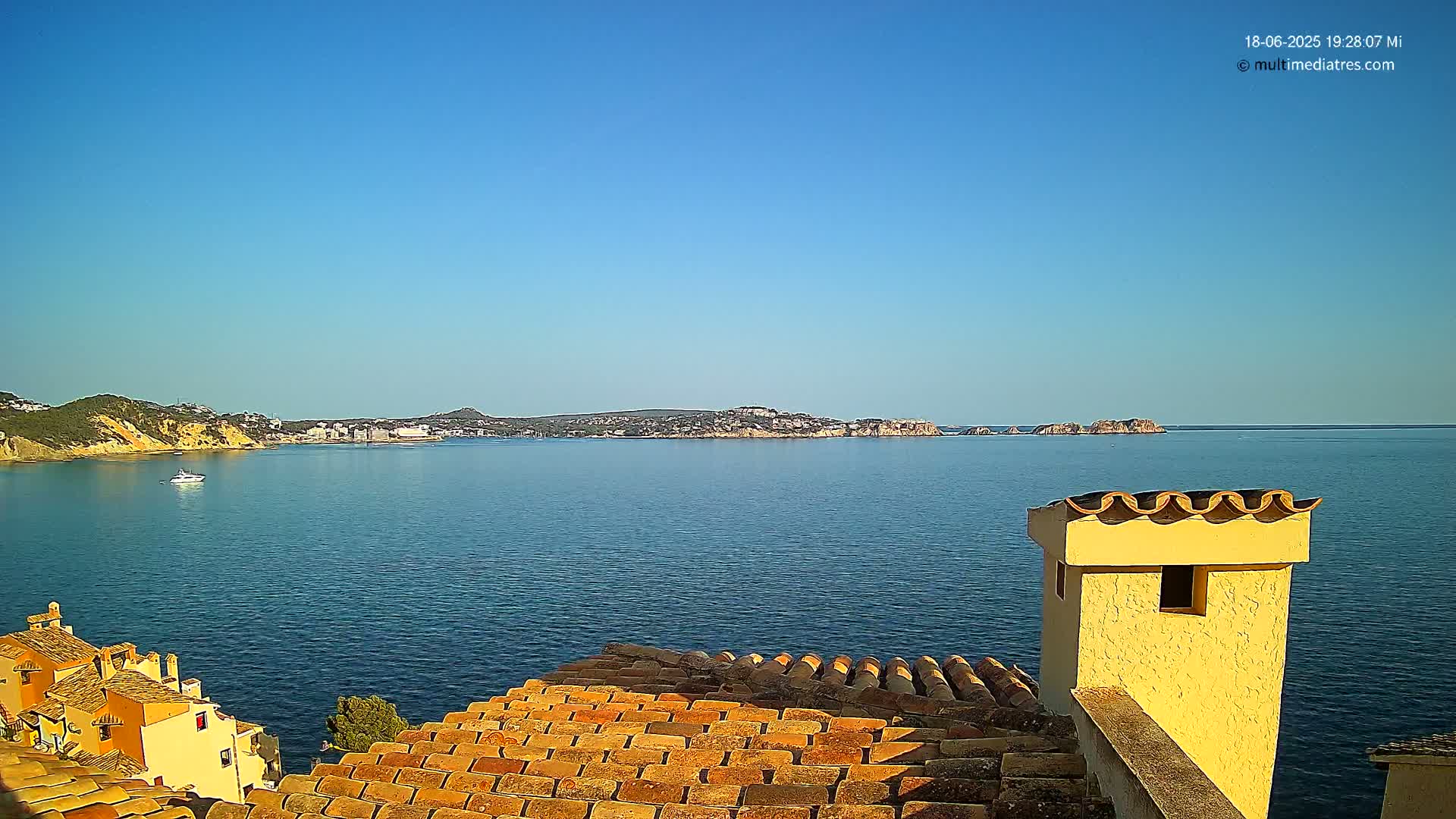 A clear blue sky overlooks a calm blue sea with a distant coastline and a yellow chimney on a terracotta roof in the foreground.