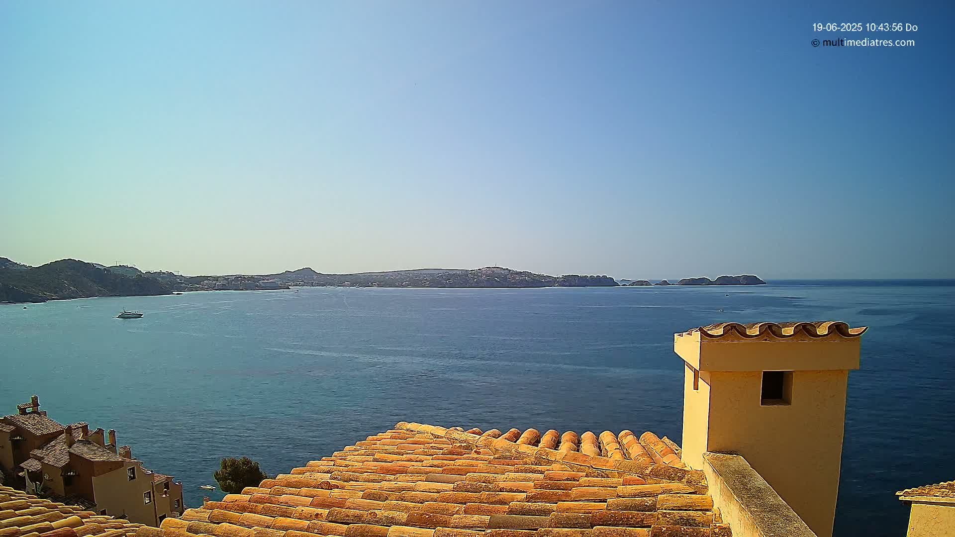 A clear, sunny day overlooks a calm blue sea from a terracotta roof with a chimney, showing a distant coastline.