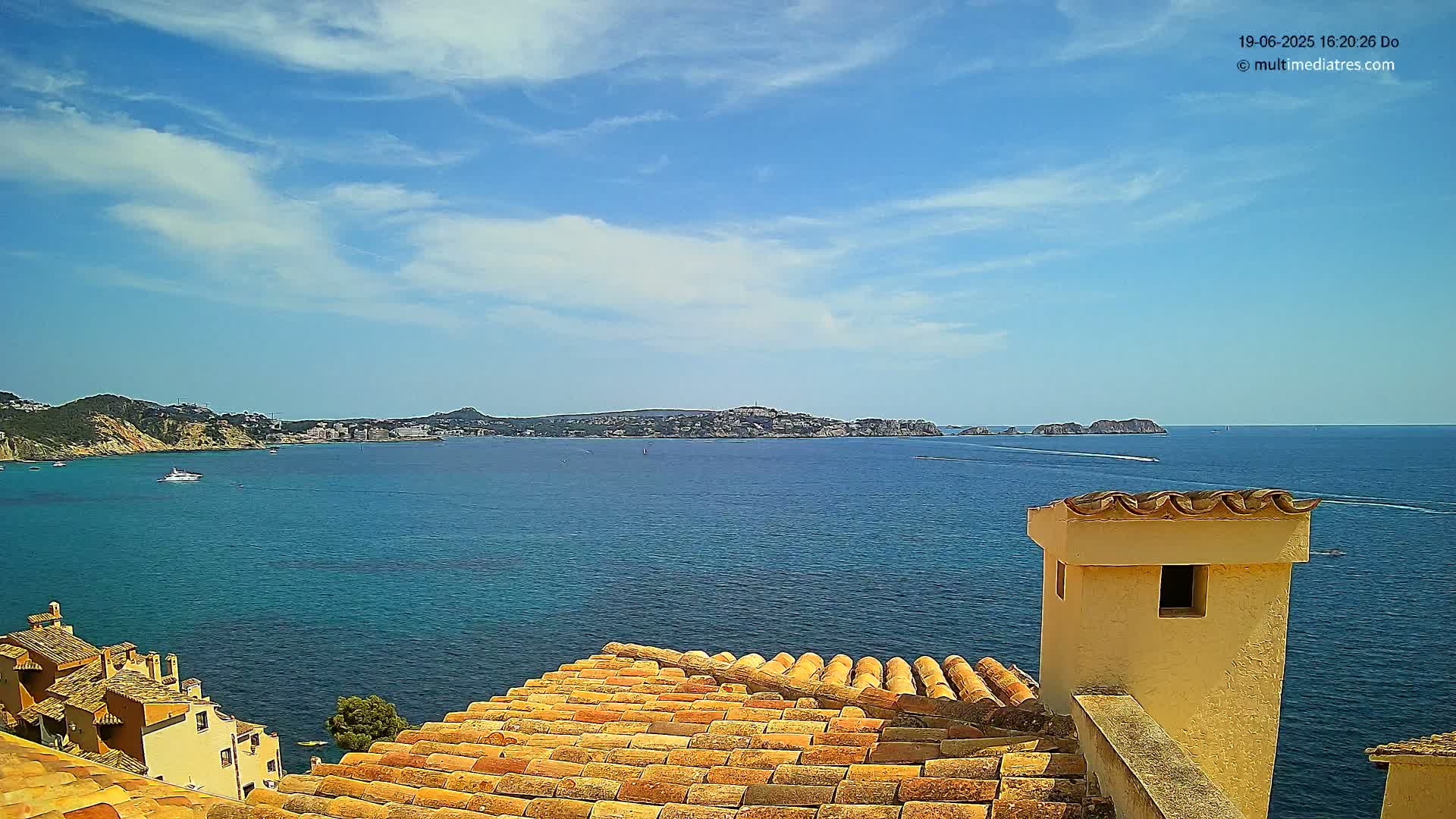 A terracotta roof overlooks a calm blue sea with a distant town and islands under a mostly sunny sky.