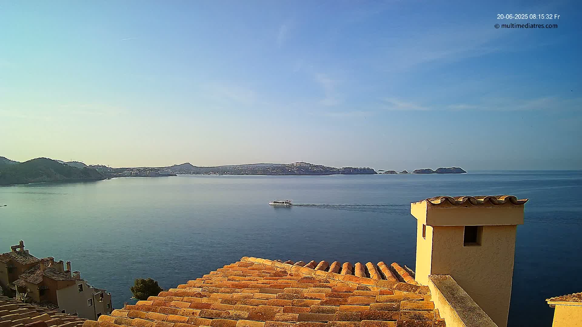 A coastal scene on a clear, sunny day shows a tiled rooftop overlooking a calm blue sea with a small boat and distant hills.