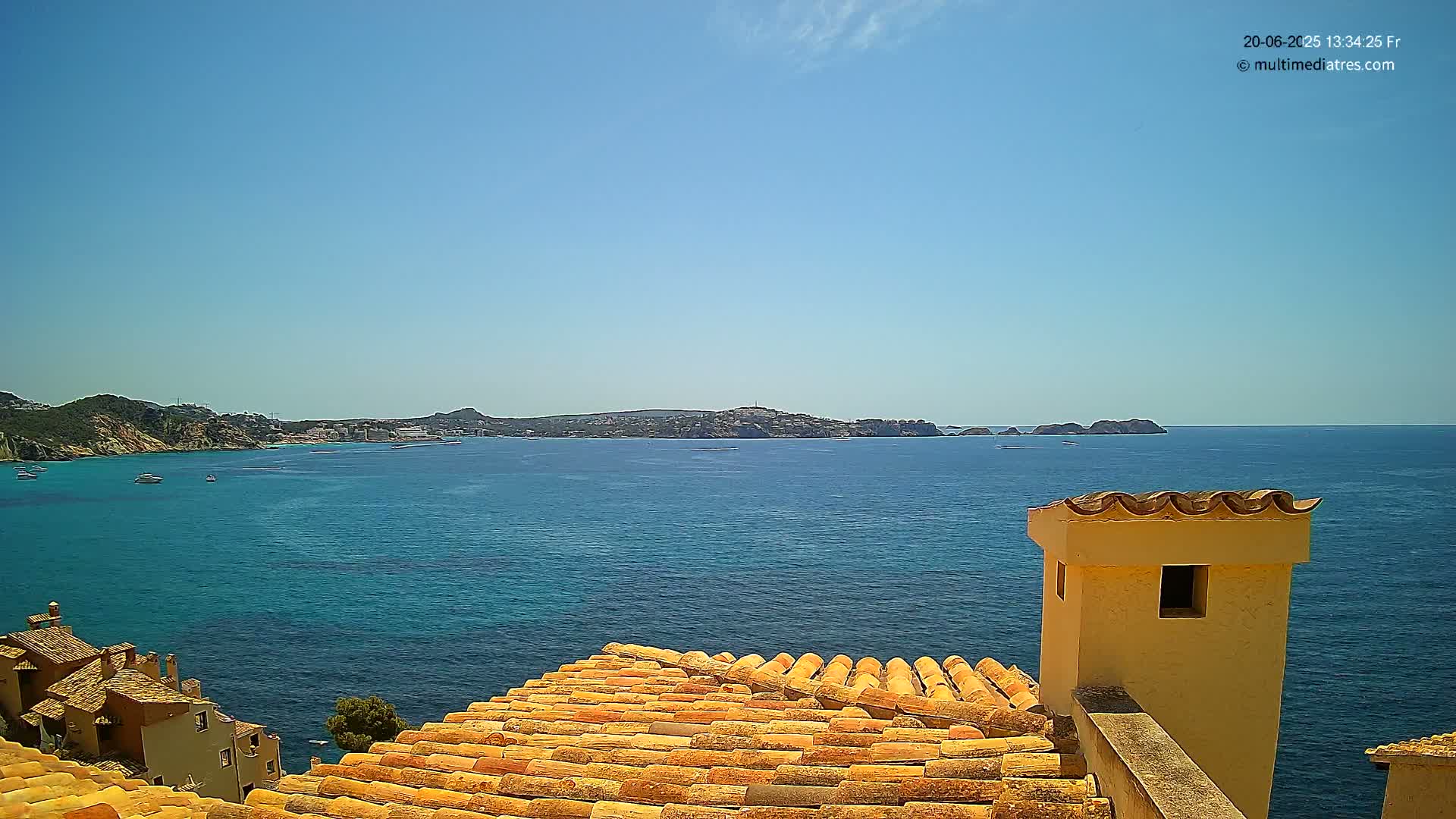 A terracotta roof overlooks a calm, blue sea on a sunny day, with distant hills and a few boats visible.