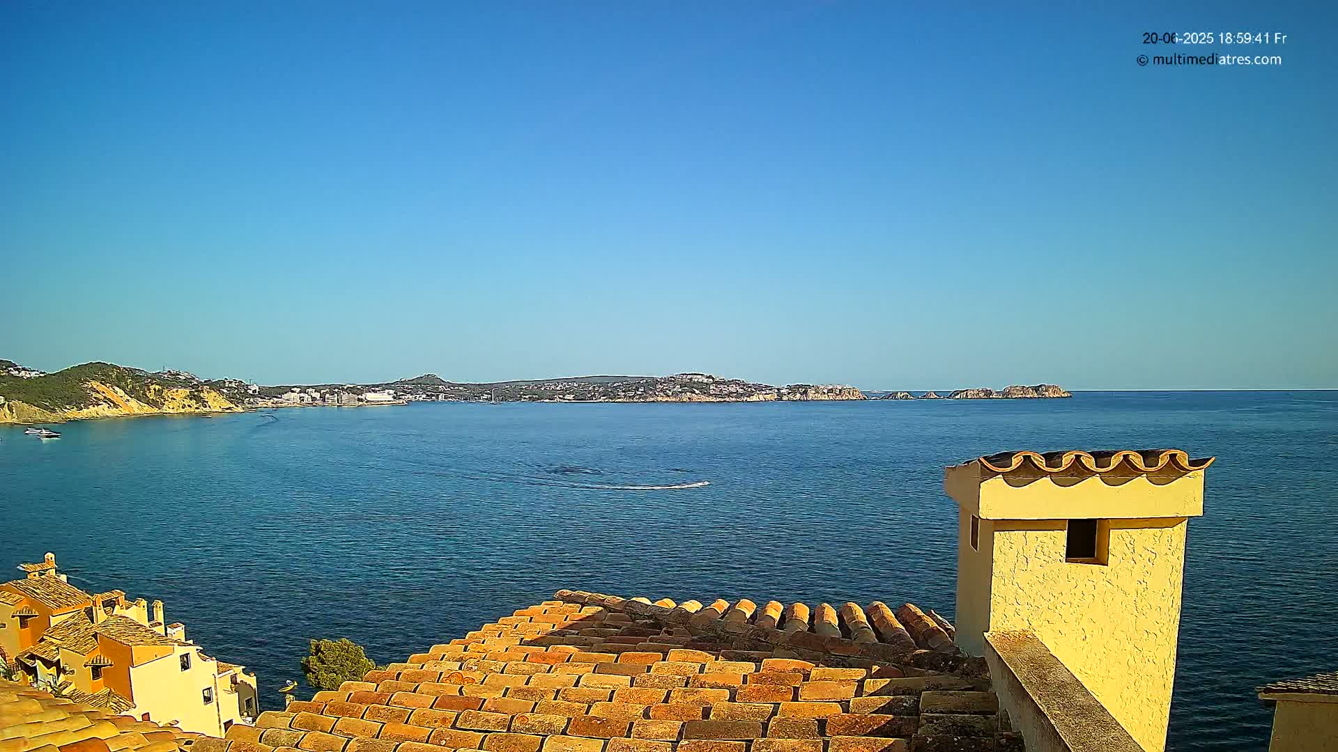 A clear, sunny day reveals a tranquil blue sea, extending to a distant coastline dotted with buildings and hills, viewed from a rooftop with terracotta tiles and a light yellow chimney.