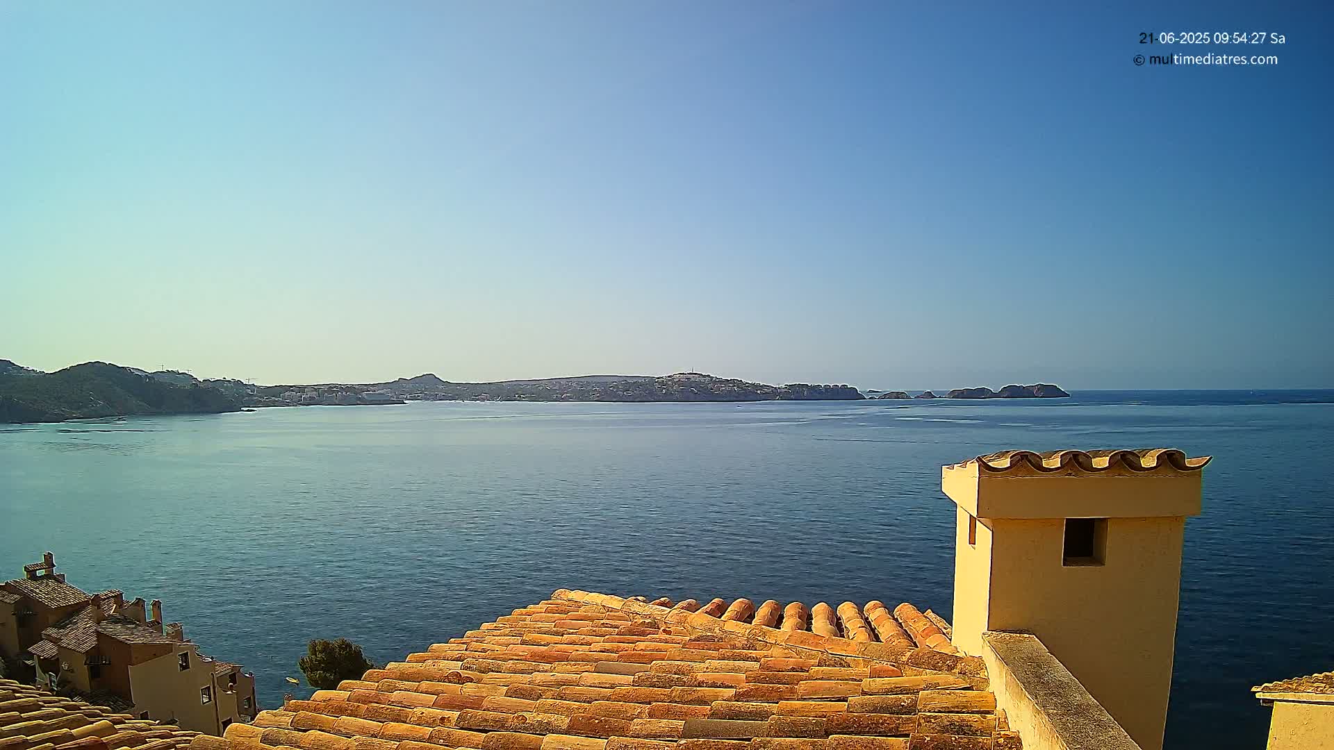 A clear, sunny day overlooks a calm blue sea from a terracotta roof with a chimney, showing a distant coastline.